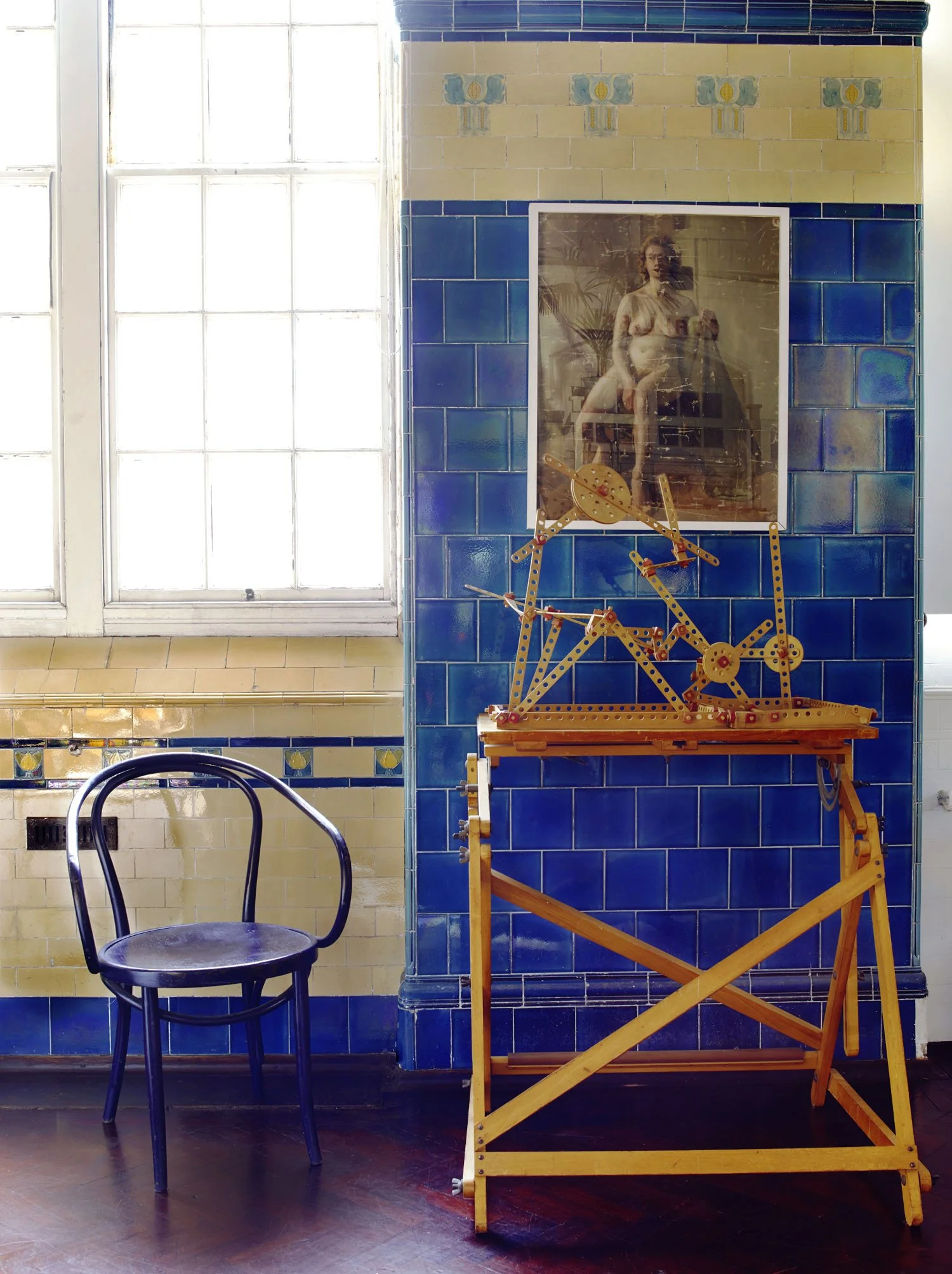 Interior scene with a large window, a black chair, a framed art piece of a nude woman, and a wooden mechanical model on a folding stand, all on a tiled wall with blue and yellow tiles.