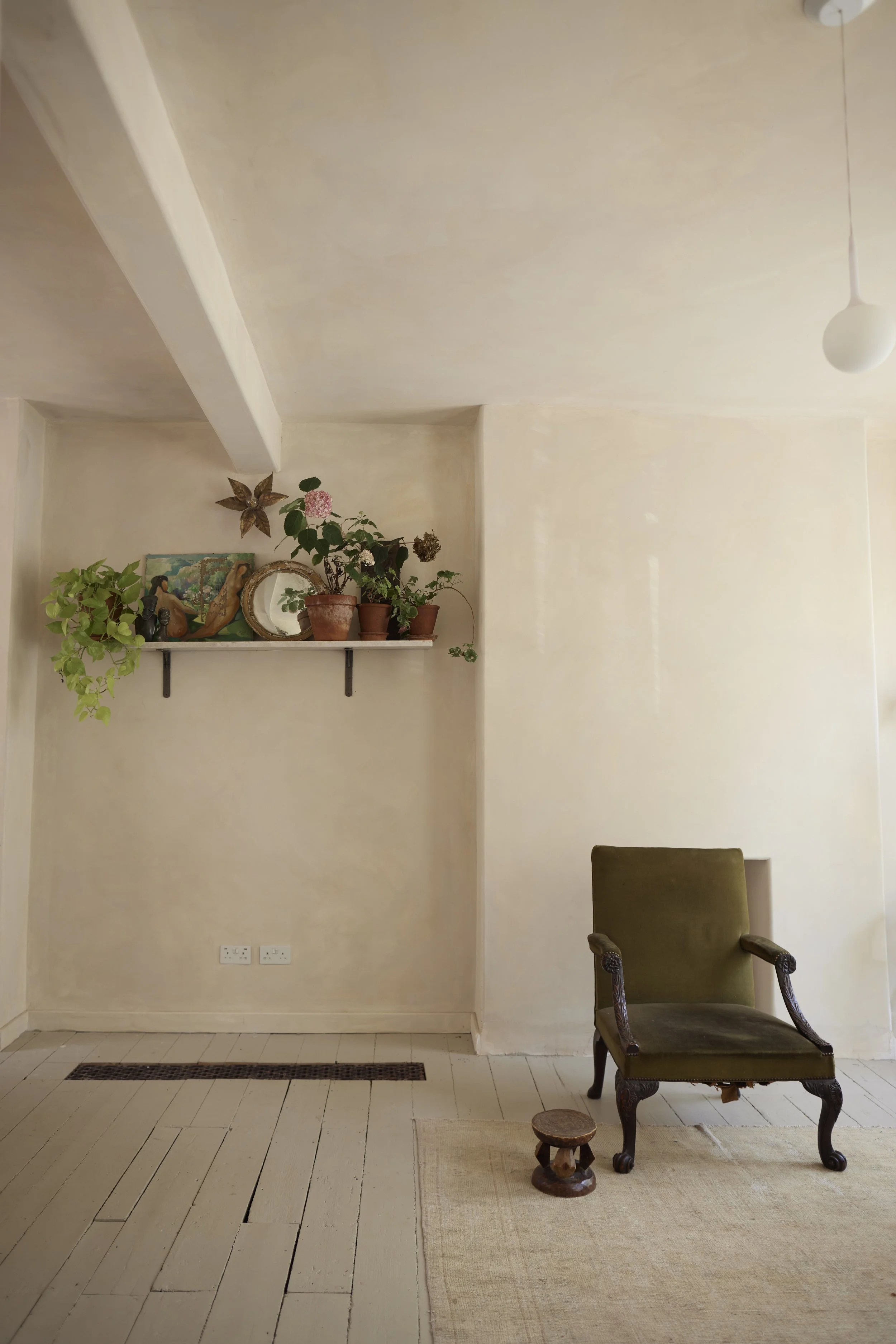 A beige living room corner with a wooden chair, small wooden stool, beige rug, a wall shelf with potted plants and decorative items, and a ceiling light fixture.