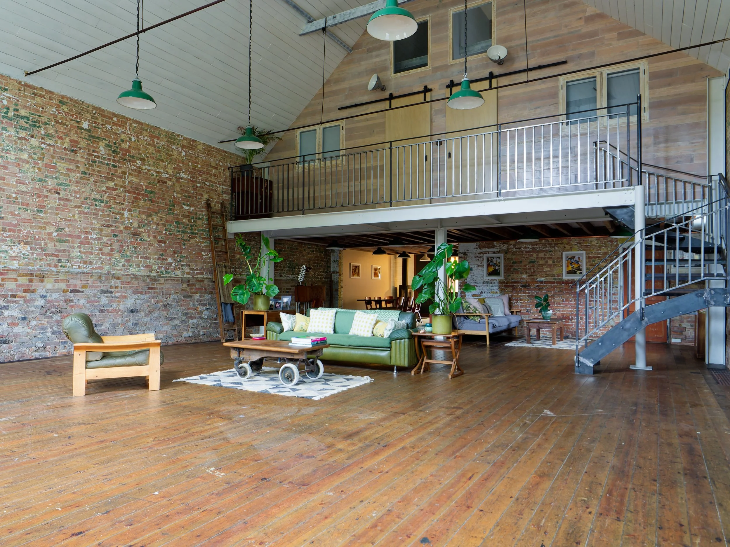 Open living space with exposed brick wall, wooden flooring, and an industrial metal staircase leading to a loft. The room is furnished with a green sofa, a wooden armchair, and various plants. A dining area can be seen in the background.