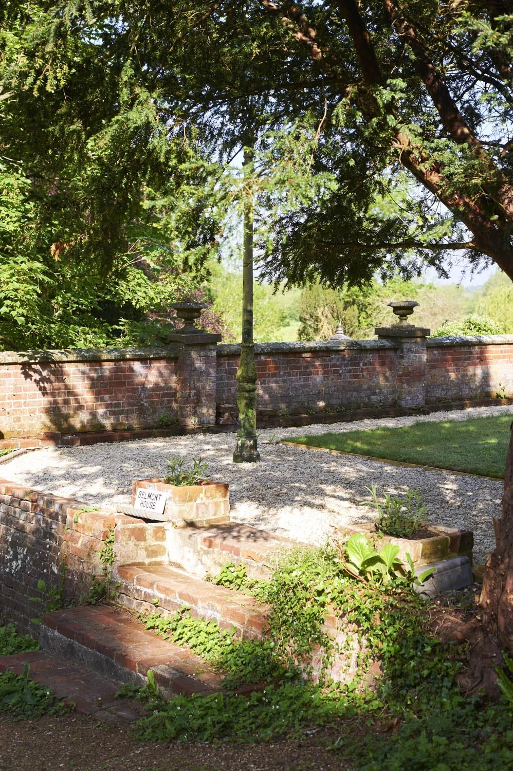 A small brick outdoor garden area with a low brick wall, a stone lantern on a pedestal, and a sign reading 'Belmont House'. The garden has plants, a gravel path, and is shaded by trees.
