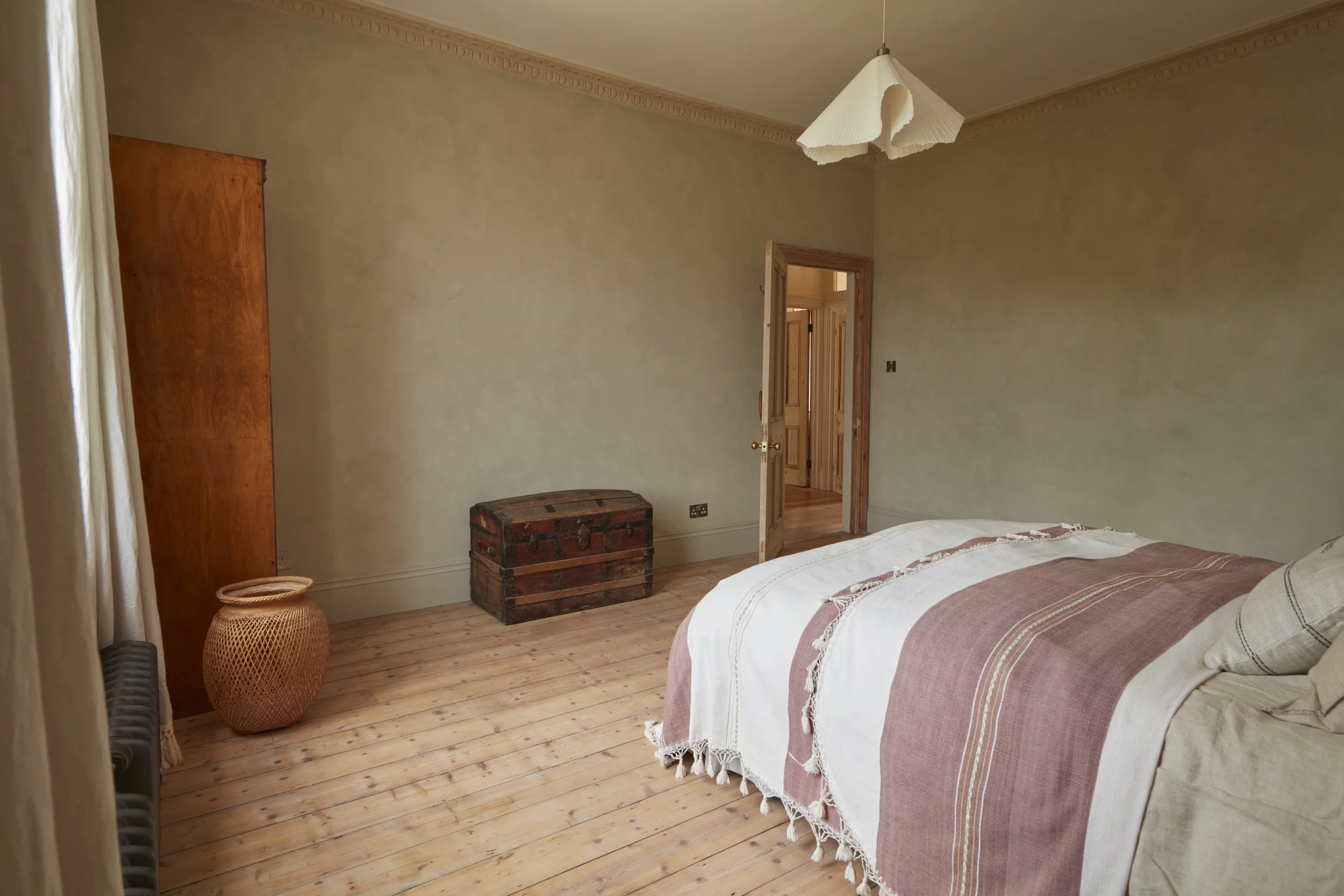 A bedroom with wooden floors, a bed with a white and pink bedspread, a wooden wardrobe, a woven basket, a vintage trunk, and an open door leading to a hallway. The room has beige walls and a ceiling light fixture.