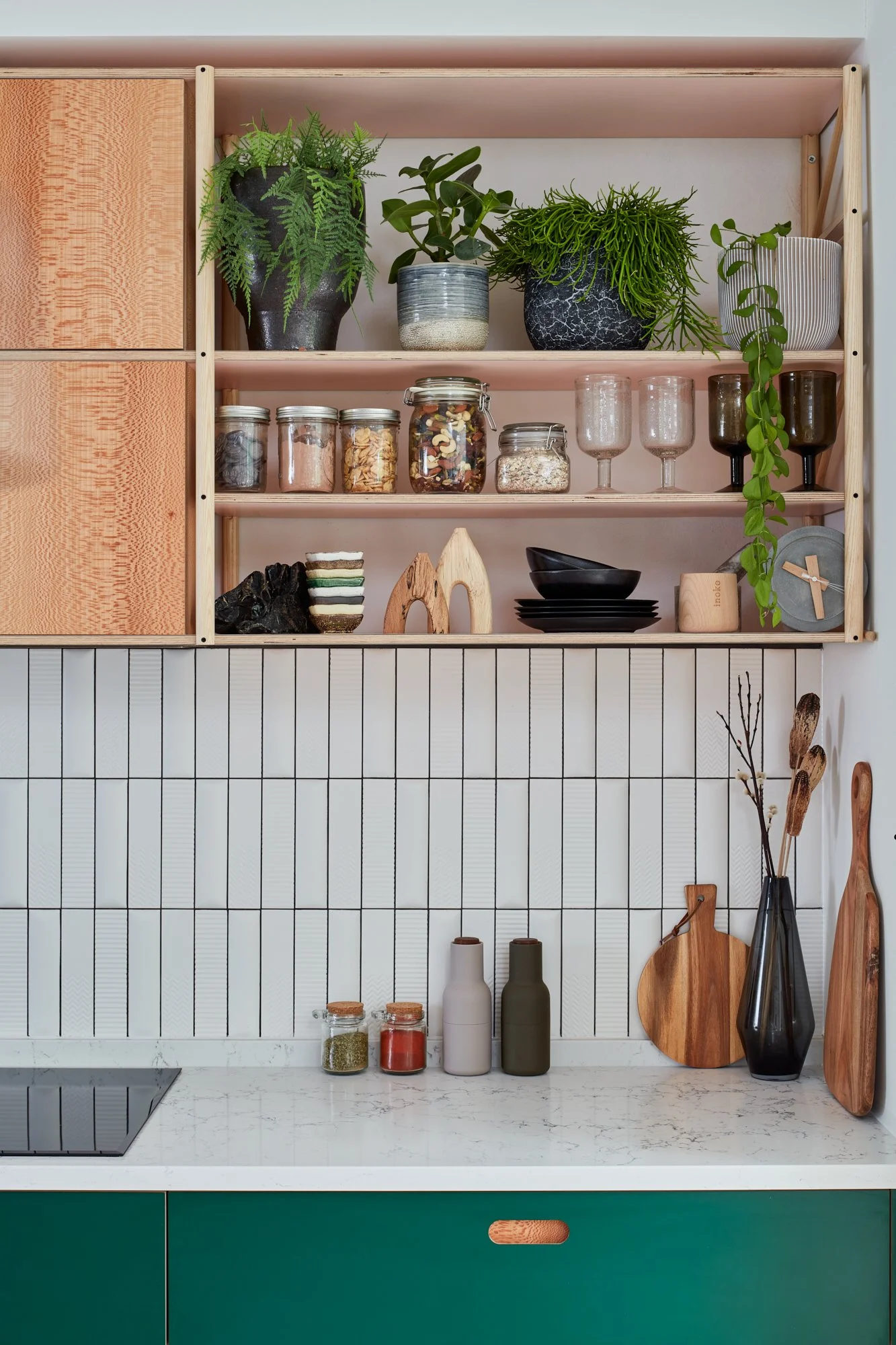 Open wooden shelf with plants, jars, and dishes above a white tiled kitchen wall and a green kitchen cabinet with a marble countertop, decorated with vases, wooden cutting boards, and containers.