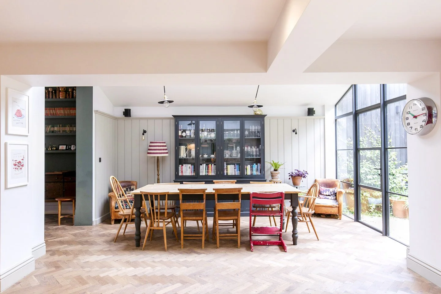 Bright dining room with a large rectangular table surrounded by wooden chairs, a black china cabinet filled with dishes, a tall floor lamp with a red and white striped shade, potted plants, large windows, and a white wall clock showing 2:10.