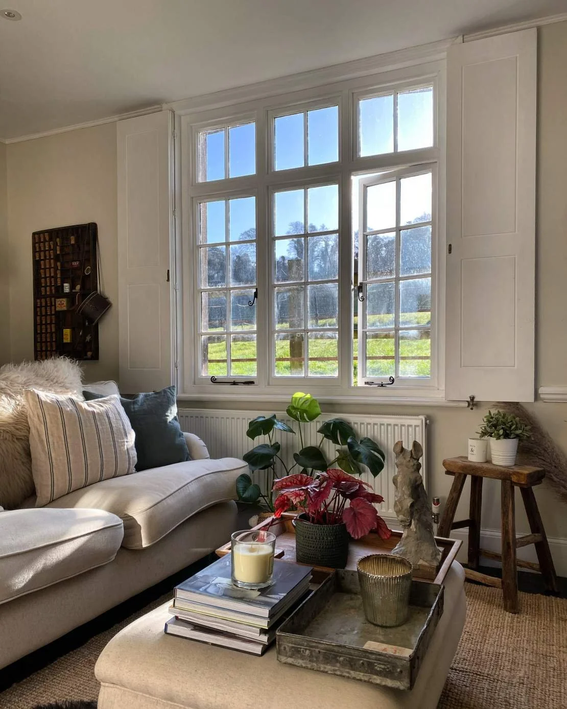 A cozy living room with a large window overlooking a green field and trees outside. The room features a beige sofa with striped and dark cushions, a coffee table with a stack of books, a candle, and a plant. There is a small wooden stool with a potte