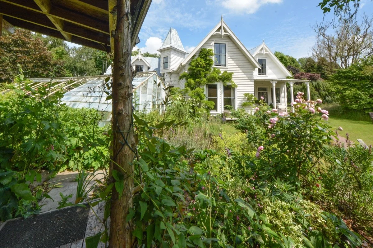 A white Victorian house with a garden and greenhouses in the front yard, surrounded by lush trees and plants on a sunny day.
