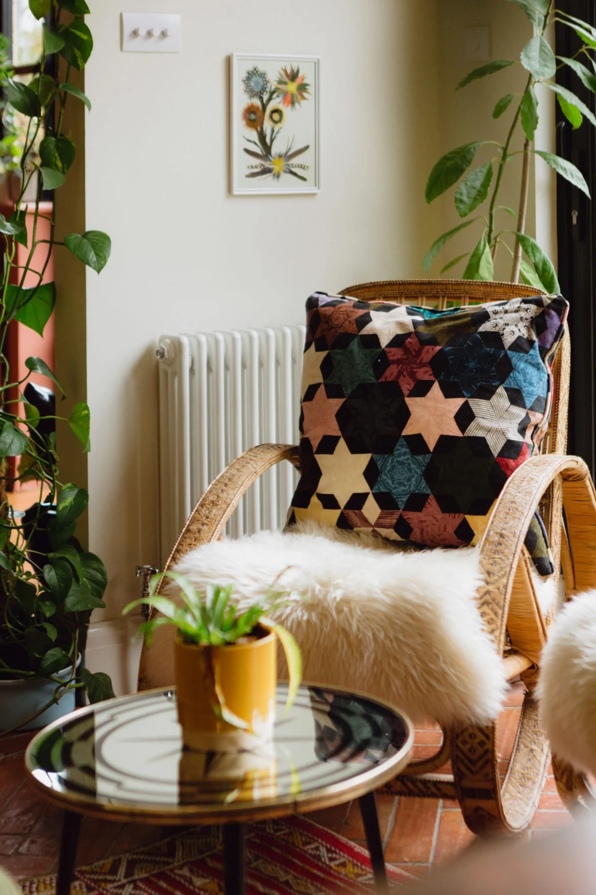 Cozy living room corner with a rattan chair draped with a colorful quilted pillow, sheepskin throw, and a small round table with a potted plant, surrounded by green plants and a wall with a framed floral artwork.