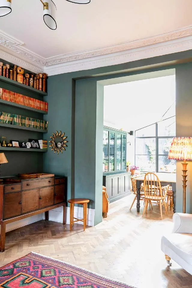 A cozy living room with greenery painted walls, wooden furniture, and a sunroom with large windows and a dining table.