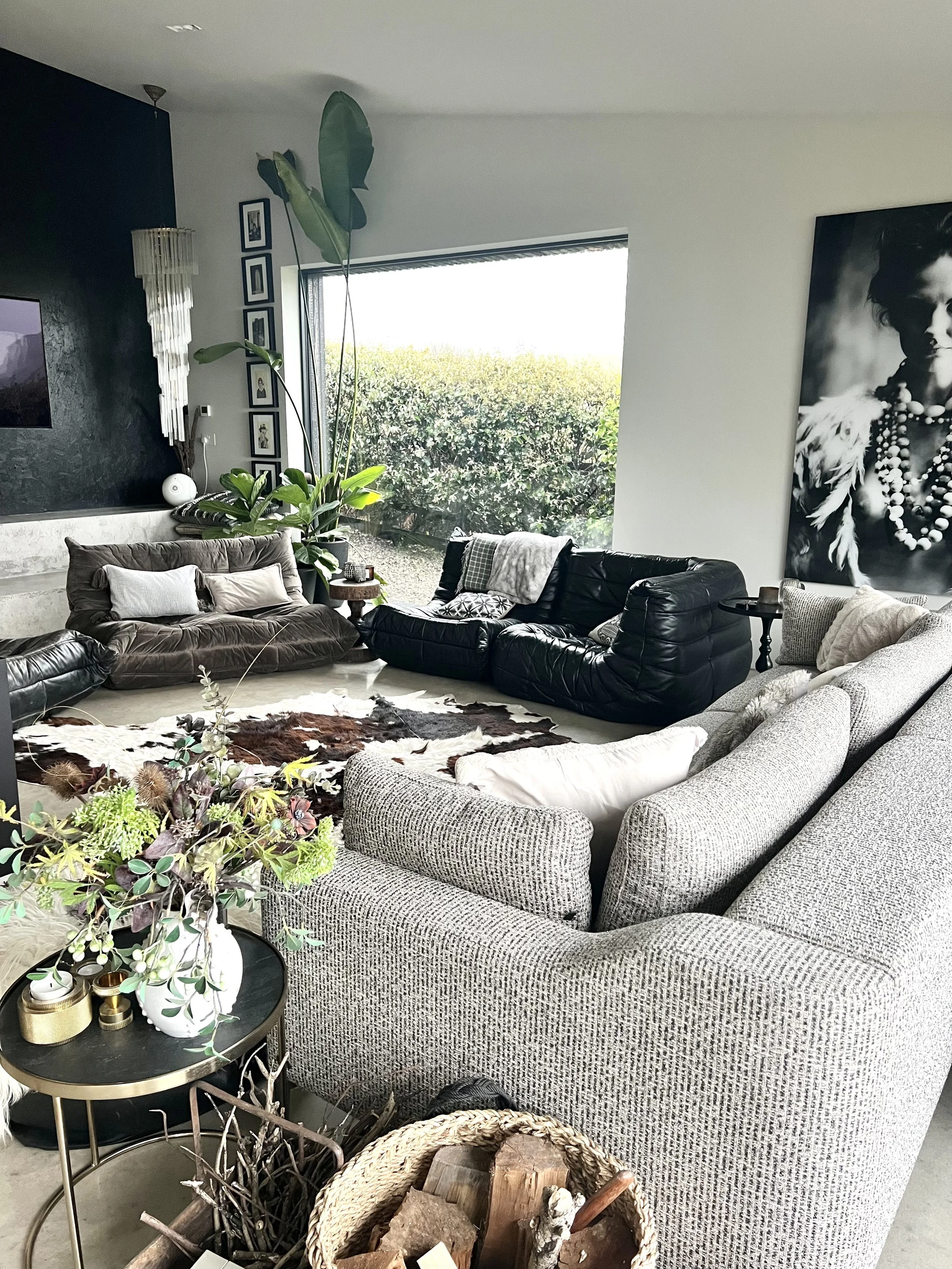 A modern living room with a large window overlooking green foliage. The room features various sofas in neutral and black colors, a cowhide rug, potted plants, framed photos on the wall, a large black-and-white portrait, and a side table with decor.