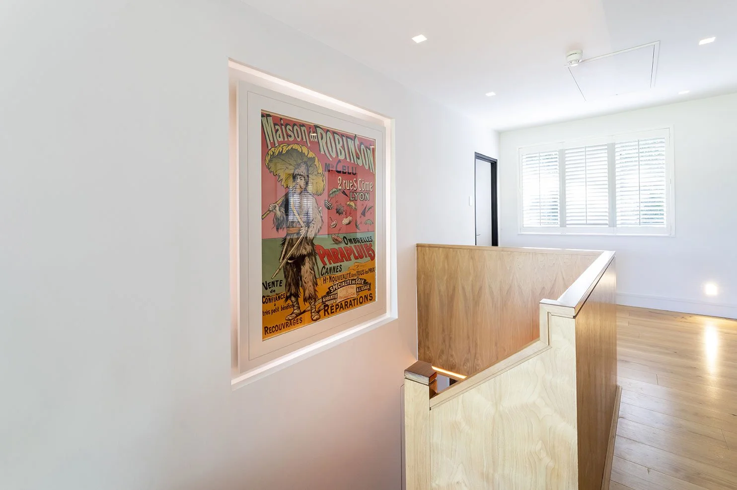 Interior of a modern, bright home featuring a colorful vintage poster on the wall near a wooden staircase with natural wood finish, overlooking a room with large window and white walls.