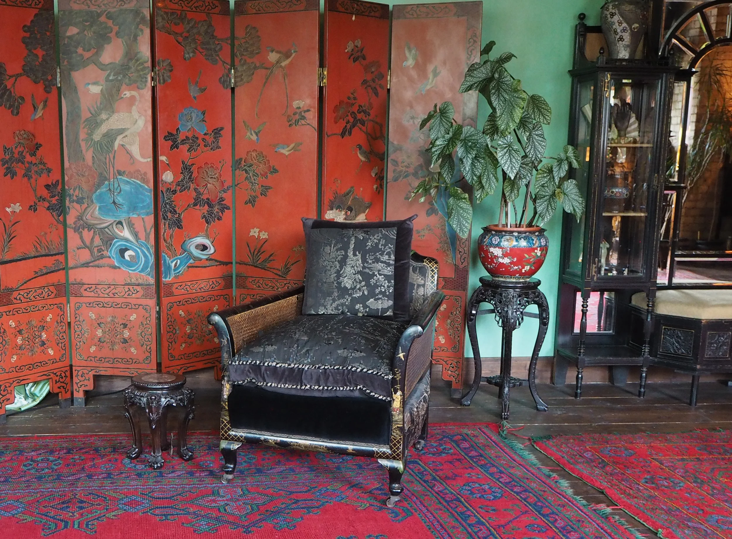 A vintage living room corner with red Asian-style folding screen decorated with birds and flowers, black patterned armchair with pillow, potted plant on ornate stand, and a dark wooden display cabinet with glass doors.