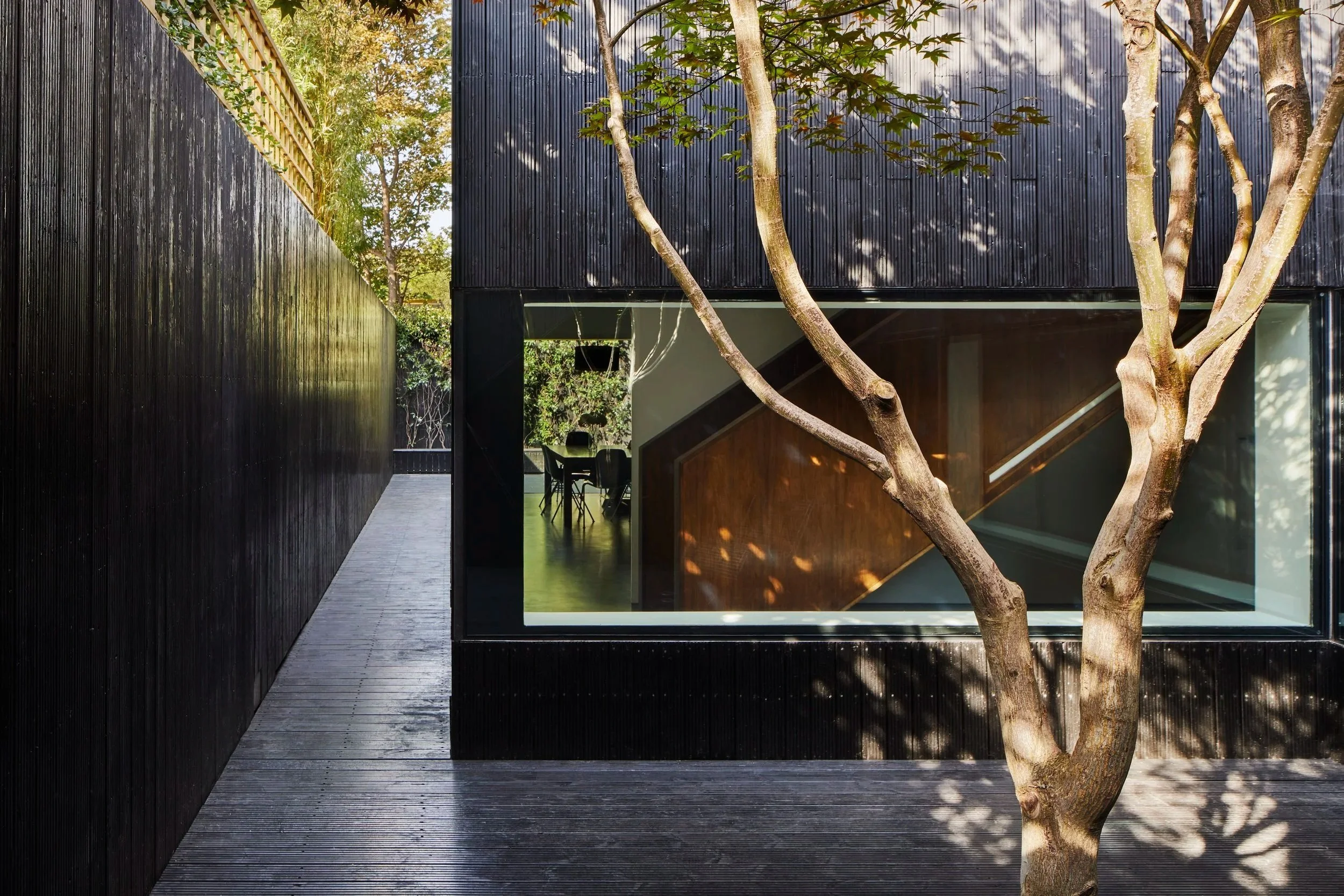 Modern building with black exterior walls, large window revealing a view of a dining area inside, and a tree in the foreground casting shadows on the wood deck.
