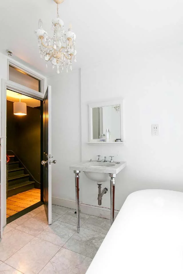 Small bathroom with white walls, a compact sink with chrome legs, a wall mirror, and a chandelier hanging from the ceiling. An open door leads to a darker hallway with stairs and a ceiling light.