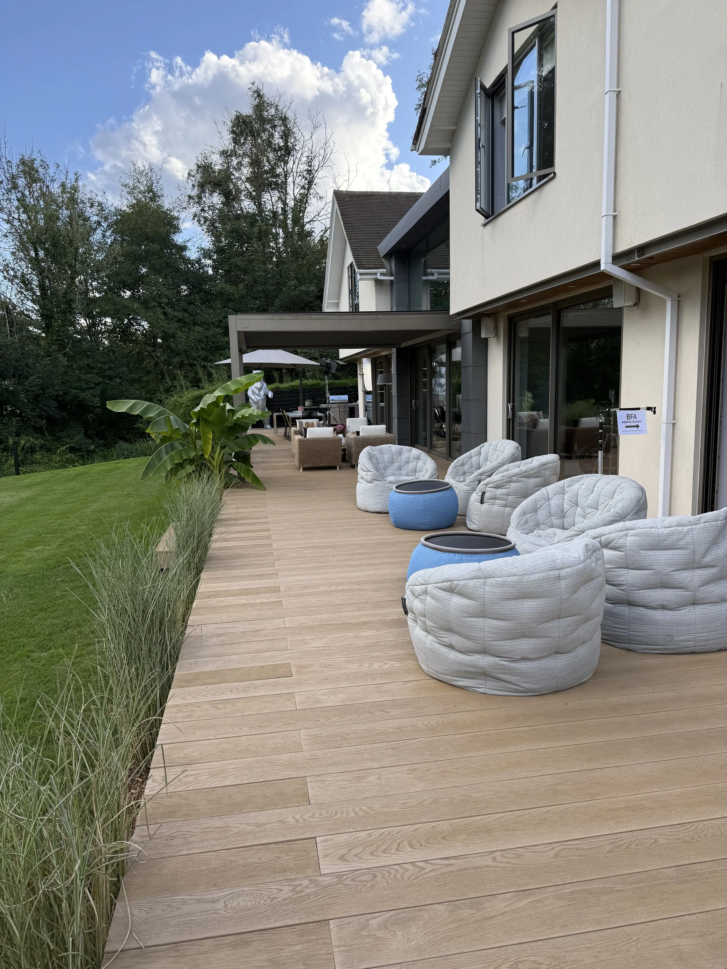 A spacious wooden deck outside a modern house with large windows, white cushioned chairs, blue side tables, plants, and outdoor furniture, with trees and a partly cloudy sky in the background.