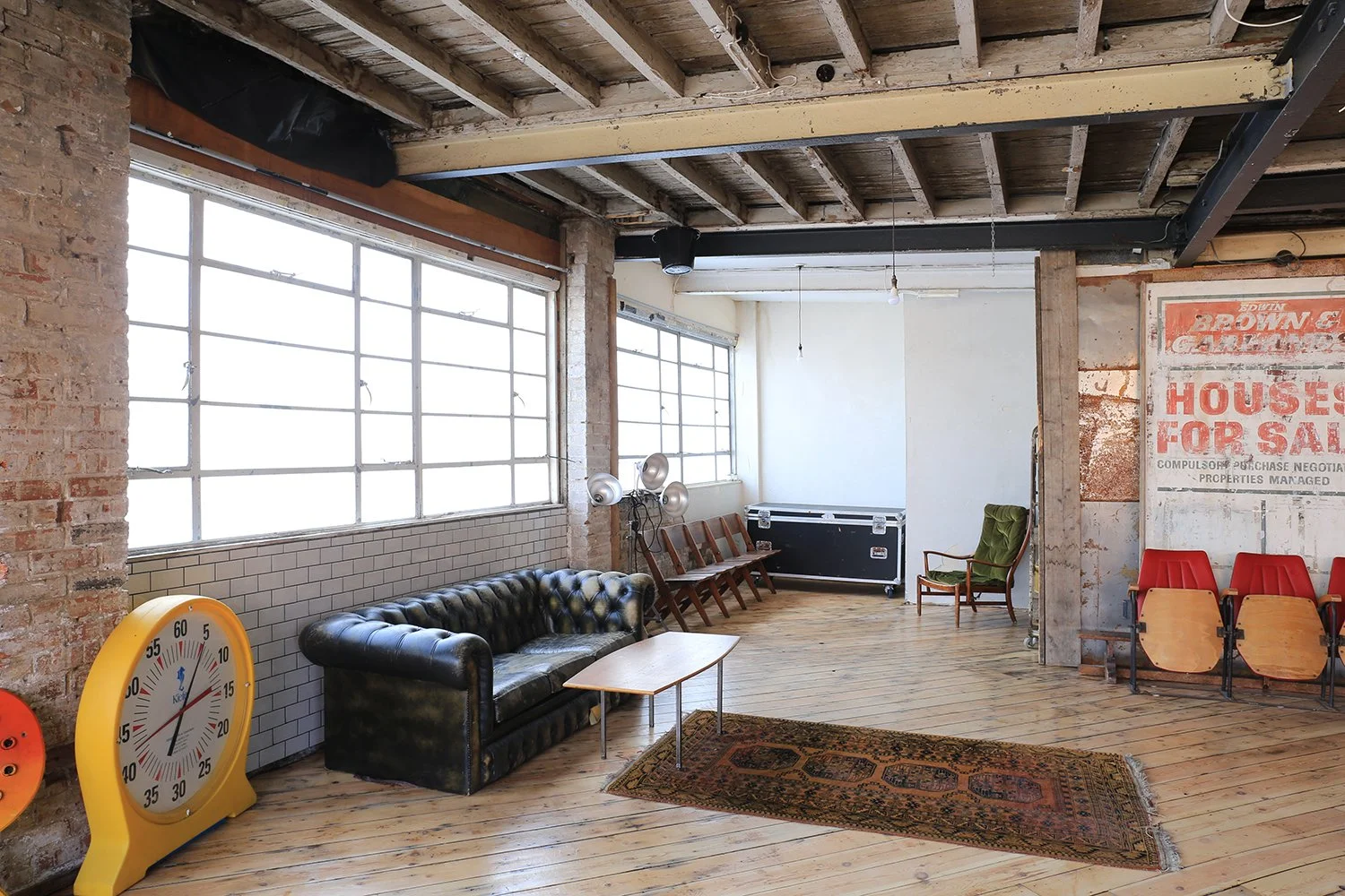 Interior of a spacious room with exposed brick walls and wooden ceiling beams, featuring large industrial-style windows, a black leather tufted sofa, a small coffee table, colorful chairs, a vintage carpet, and a large yellow clock.