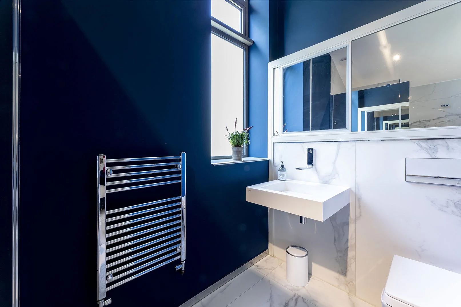 A modern bathroom with dark blue walls, a white marble wall behind a rectangular white sink, a large mirror, a window with a potted plant, a small white trash can, and a chrome towel warmer.