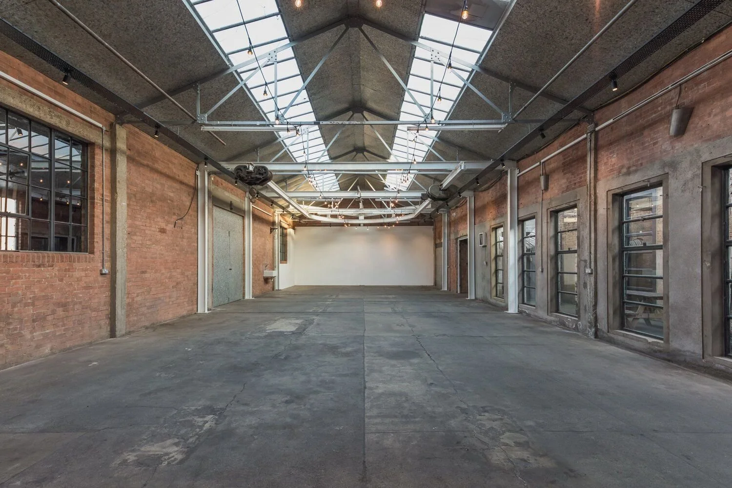 Empty industrial-style loft with large skylights, exposed brick walls, concrete floors, and multiple windows along one wall.