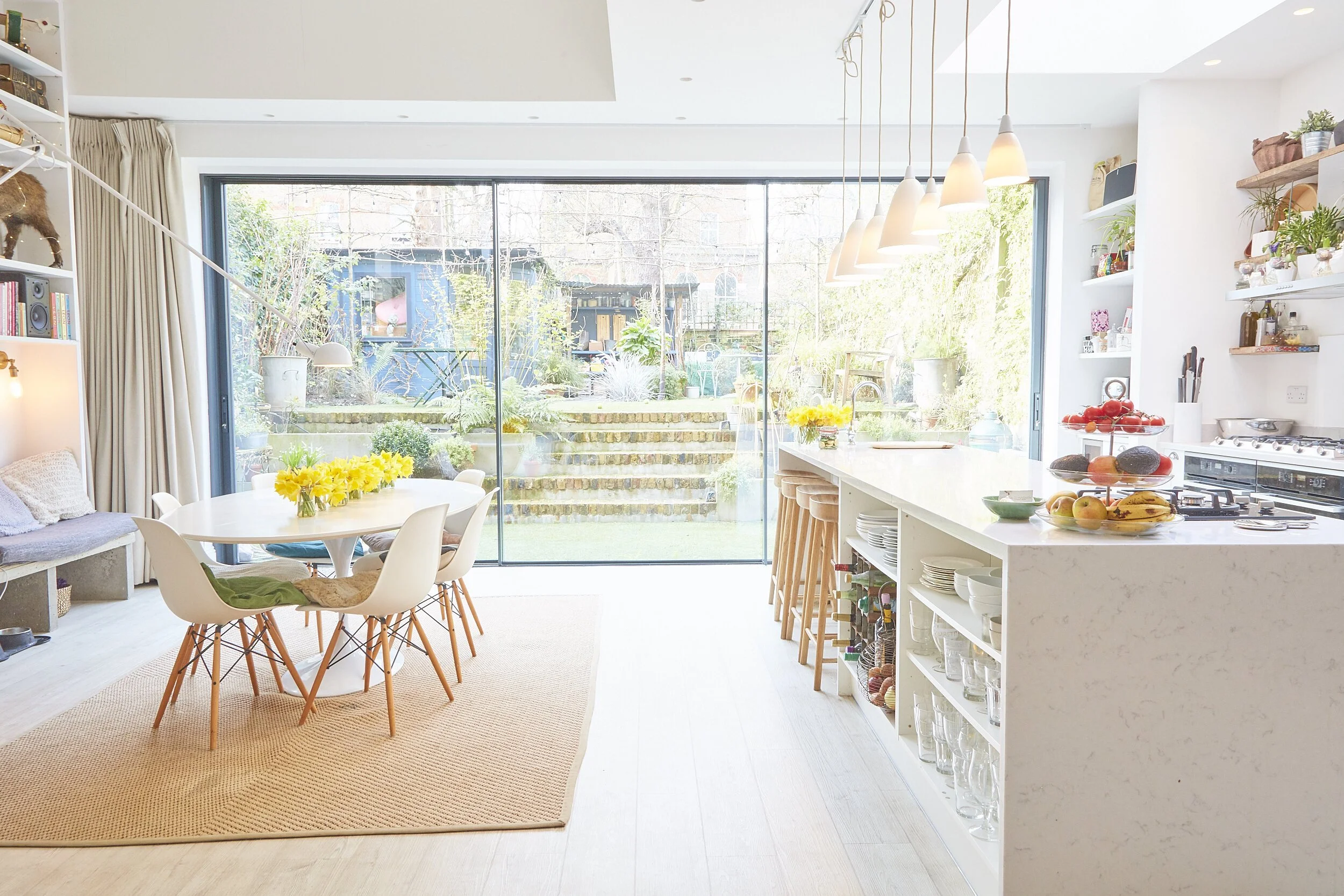 Bright, modern kitchen and dining area with a large sliding glass door opening to a backyard garden. The dining table has a yellow flower centerpiece and white chairs with wooden legs. The kitchen island has a bowl of apples, bananas, avocados, and a