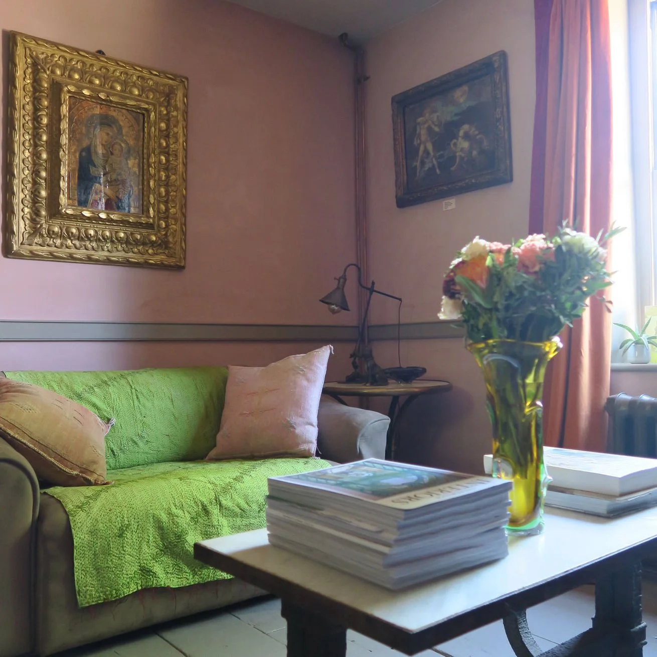 Living room with a green sofa covered with a textured blanket and pink pillows, a wooden table with a large stack of magazines and a vase of flowers, framed art on the wall, a window with curtains, and a small side table with a lamp.
