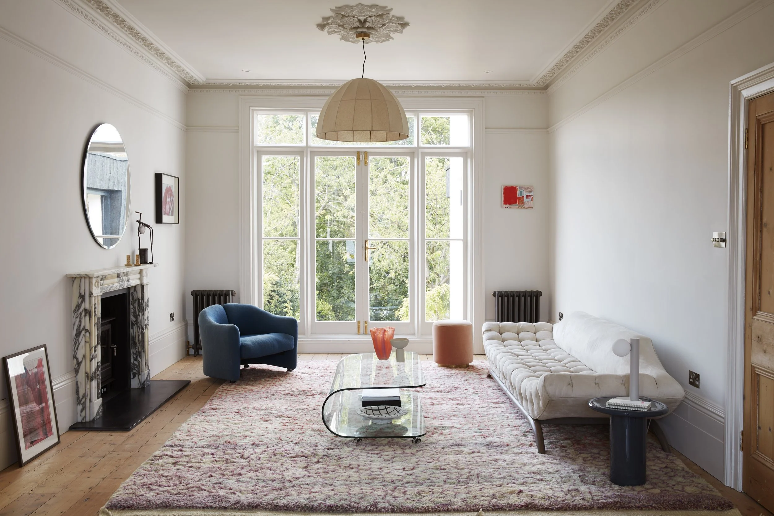 Living room with white walls and large window, featuring a blue armchair, a cream tufted sofa, a glass coffee table, a black side table with a lamp, colorful artwork, a decorative marble fireplace, and wooden flooring with a patterned rug.