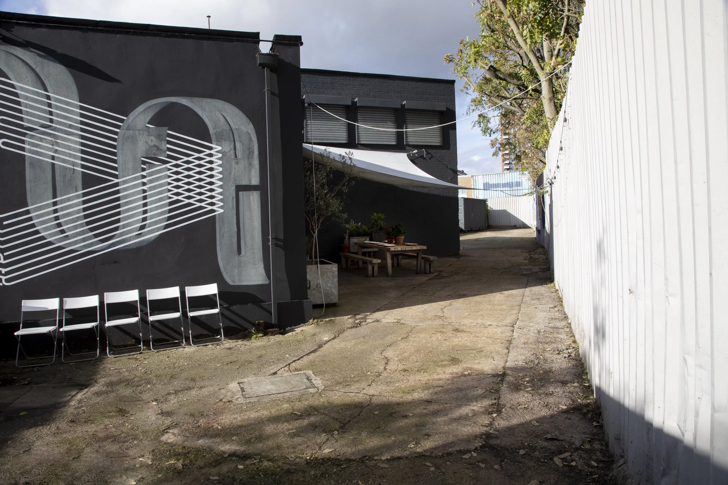 An alleyway with a black wall on the left featuring a large white geometric mural, four white chairs lined up against the wall, potted plants on a wooden table, and a white fence on the right. The sky is partly cloudy with some trees visible at the e