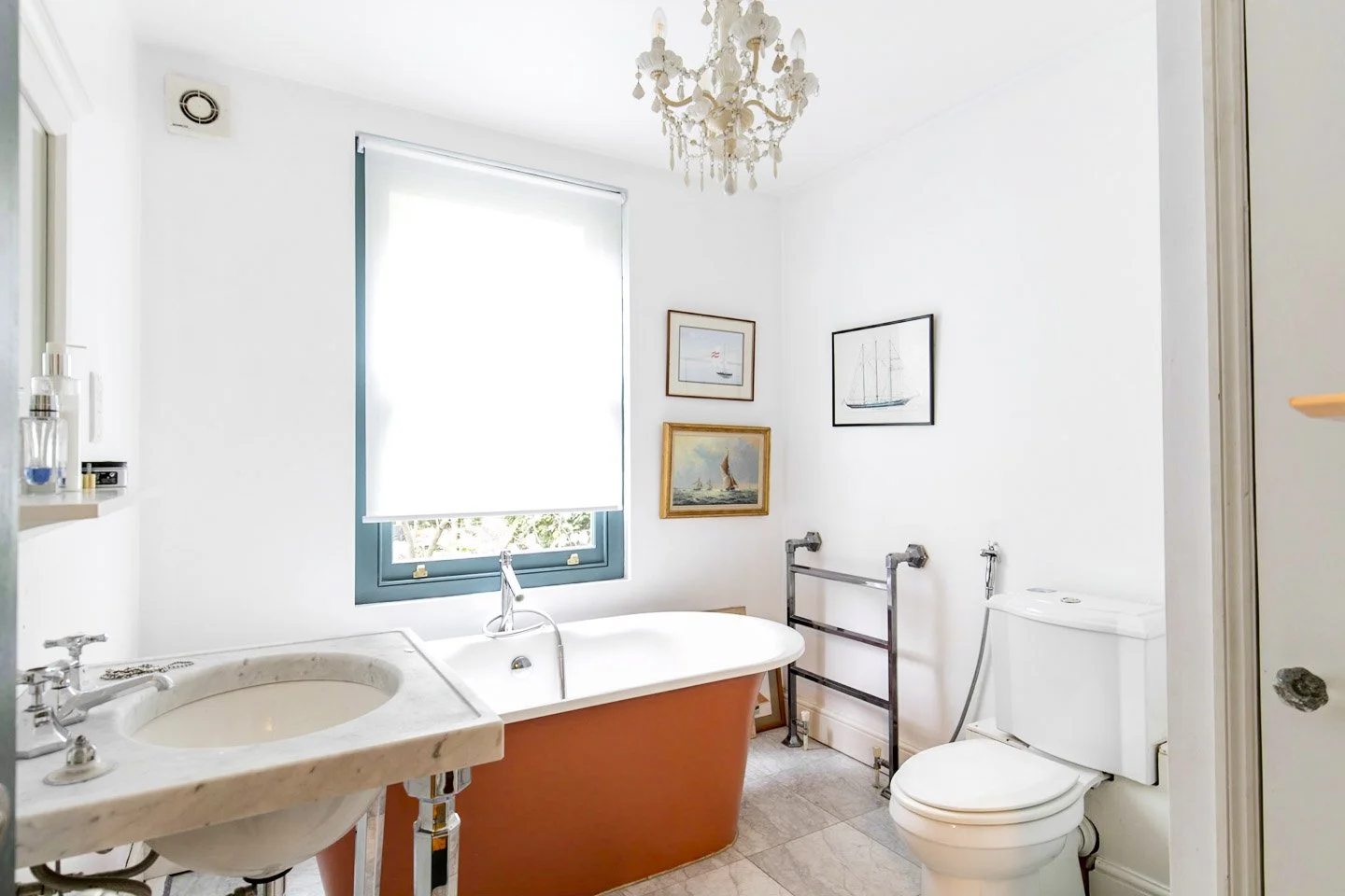 Bathroom with a white bathtub, a marble sink, a toilet, a towel rack, framed pictures of sailboats on the wall, and a chandelier. Natural light coming through a window with a closed white shade.