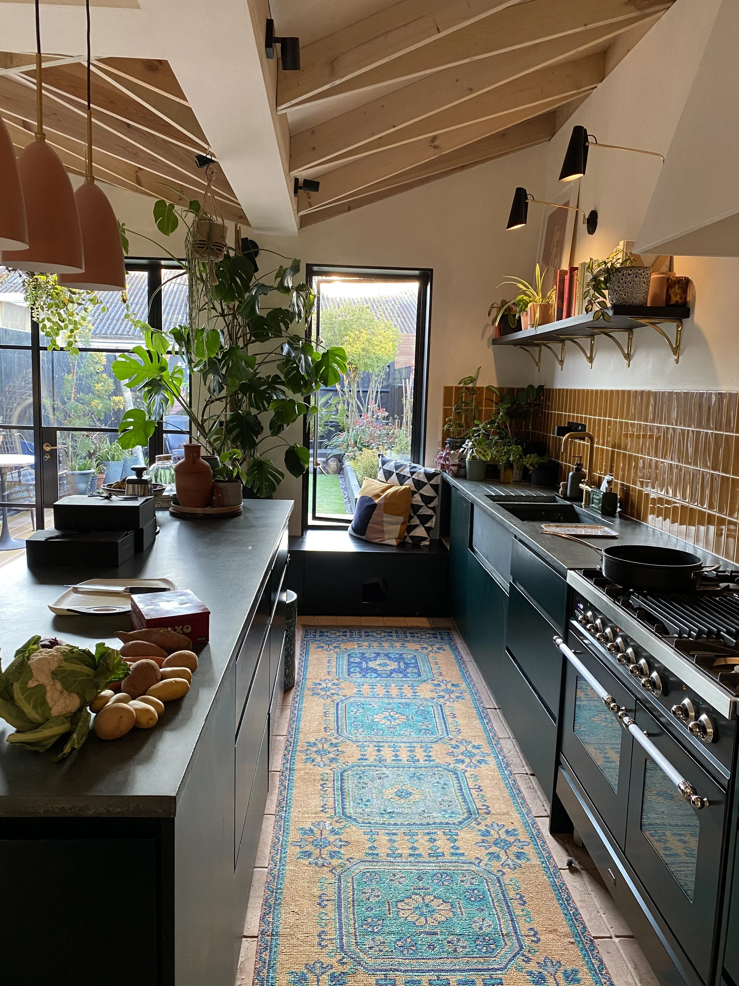 Kitchen with green cabinets, black countertops, orange tile backsplash, plants, and large window looking into a garden.