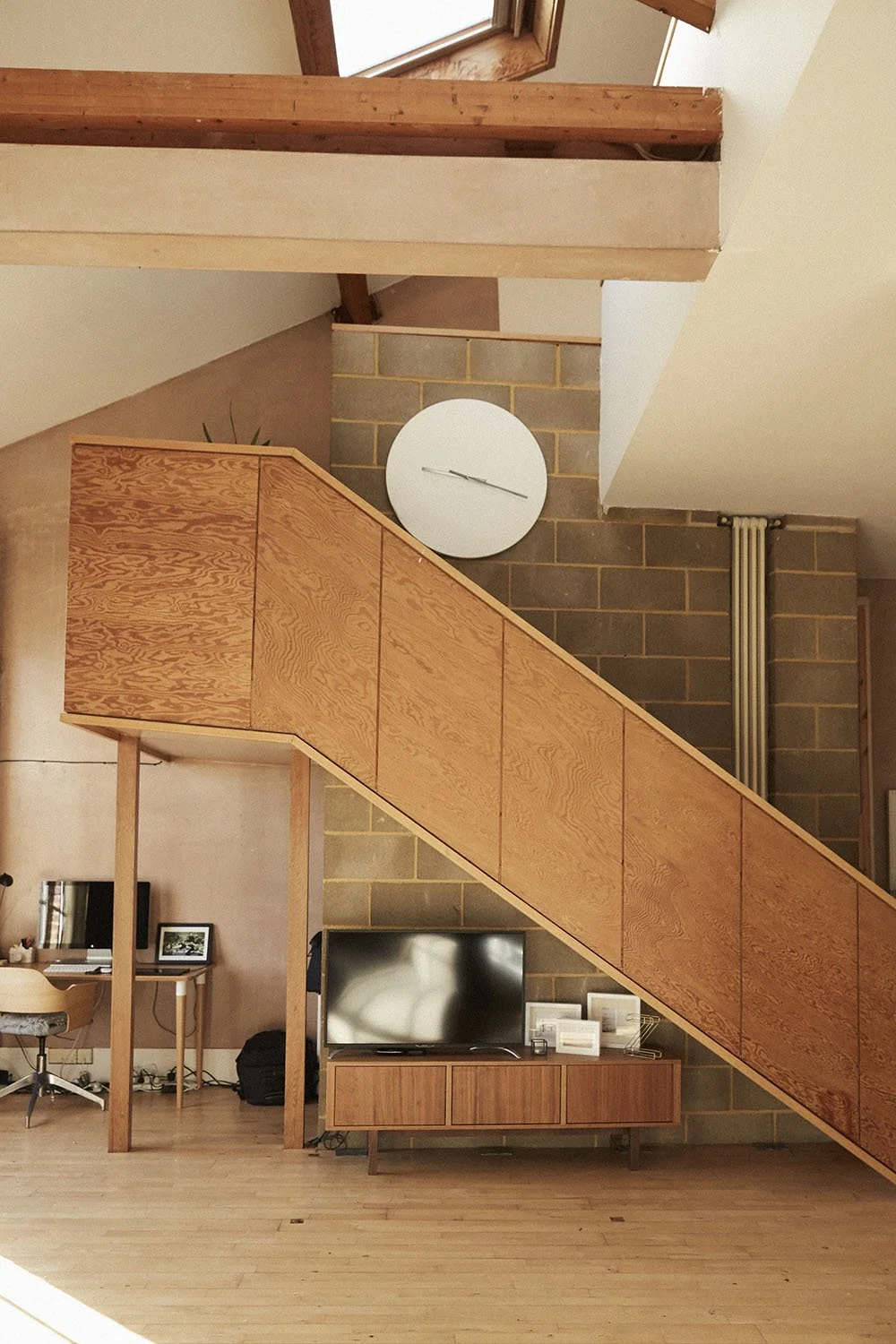 Living room with staircase featuring wooden paneling, a flat-screen TV on a wooden stand, a desk with a computer and chair, and a large round wall clock.
