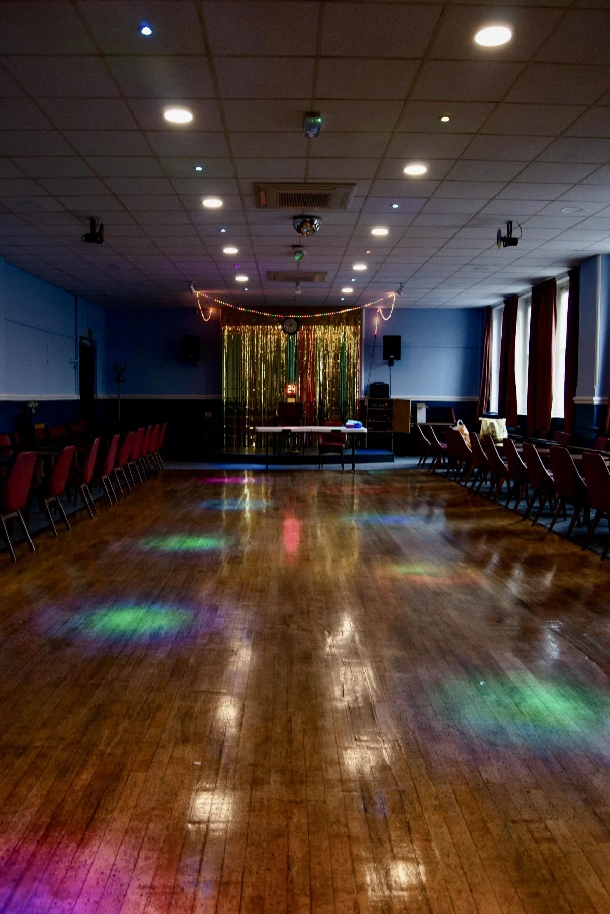 Empty dance hall with colorful stage lights and gold shimmering backdrop, wooden floor, red cushioned chairs along the walls, and a small table in the front. Smalls on the floor reflect colored lights.