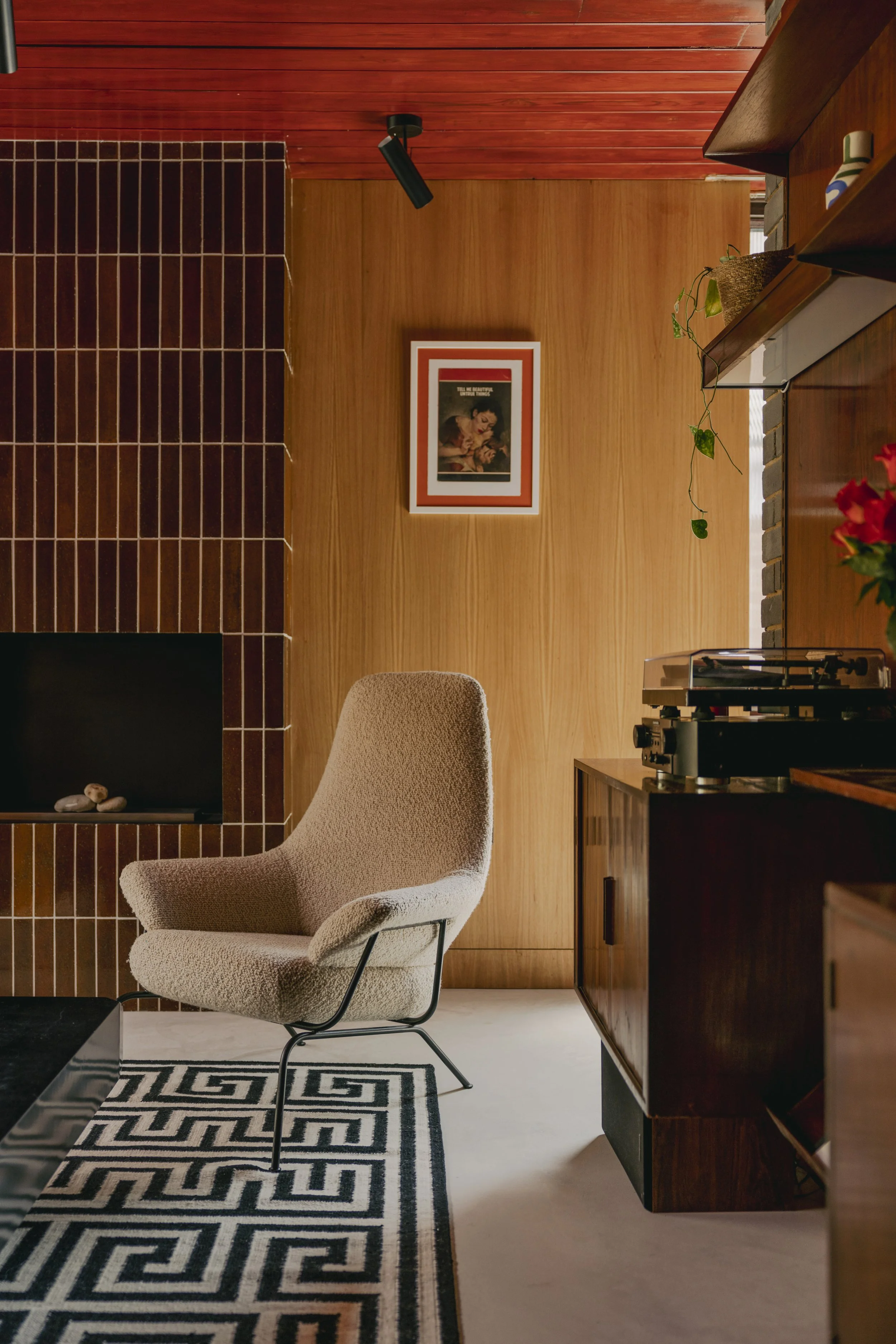 Interior of a room with a beige upholstered armchair on a black and white geometric rug, a dark wood cabinet with a record player, a brick fireplace, and a framed vintage poster on a wooden wall.