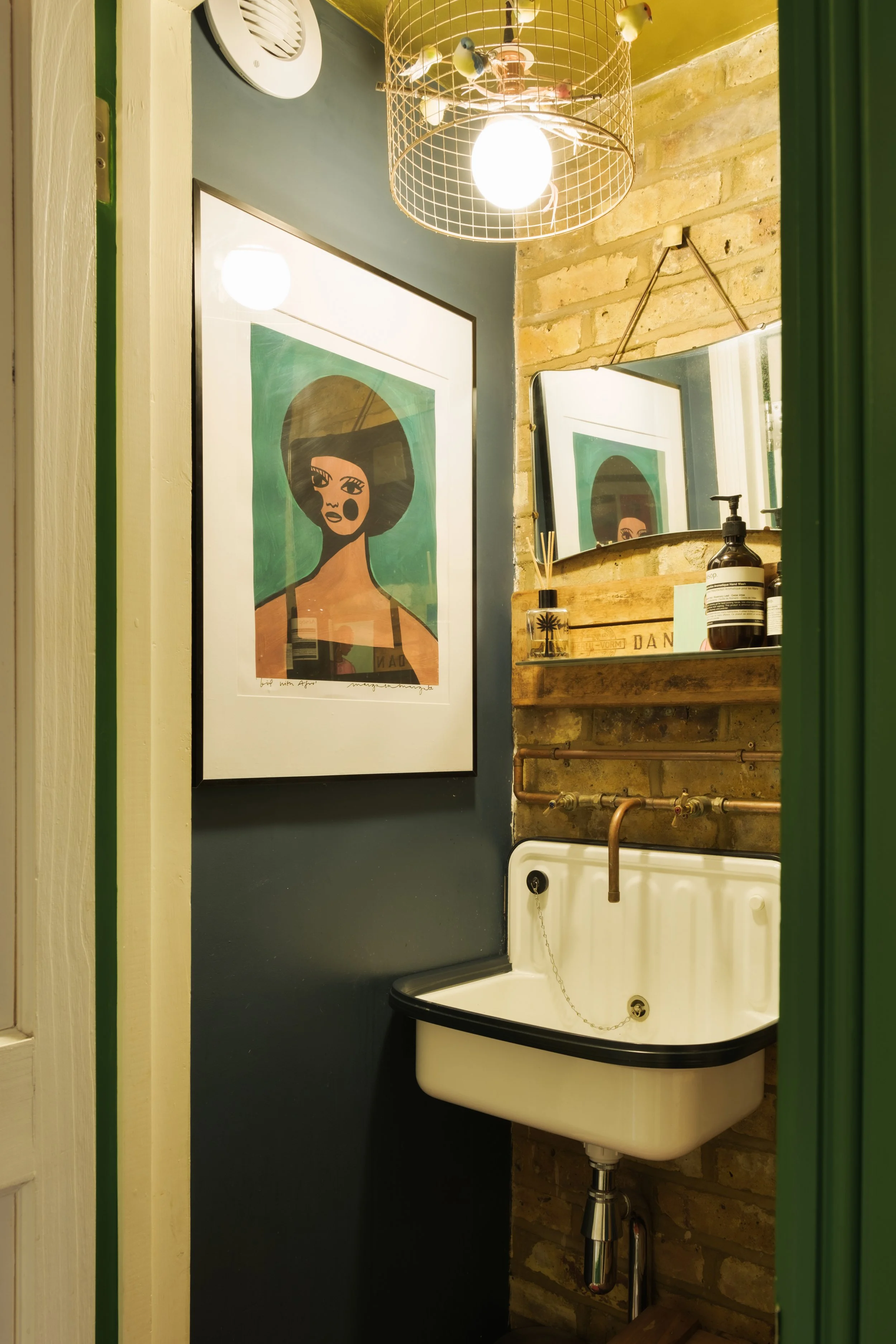 Interior of a small vintage bathroom with a white ceramic sink, a mirror, artwork of a woman with large hair, a wooden shelf with bottles and reed diffusers, a copper pipe, a brick wall, and a ceiling light fixture.