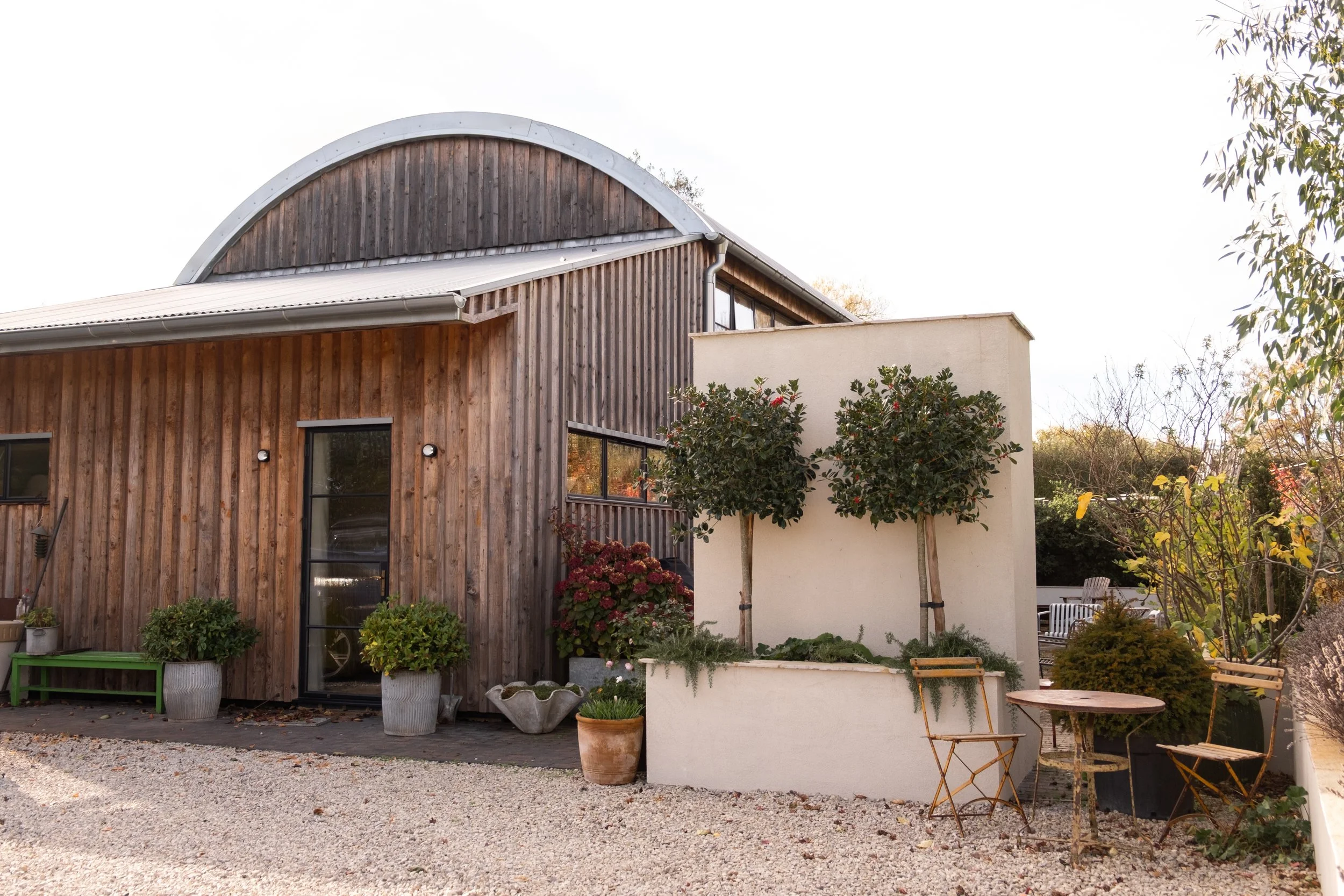 A modern house with wooden siding and a curved roof, surrounded by various potted plants and trees in a garden.