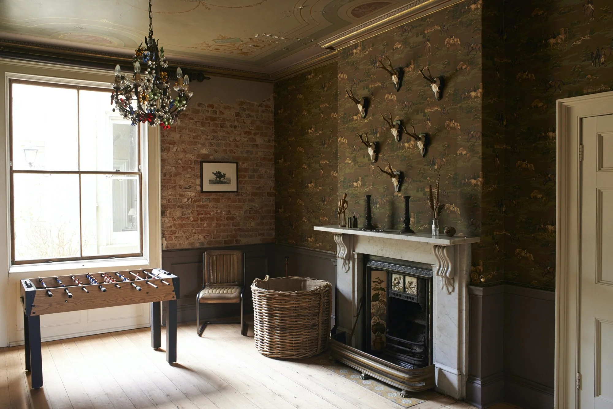 Interior of a room with a large window, a colorful chandelier, a foosball table, a leather chair, a woven basket, a fireplace with decorative objects, a wall with mounted deer antlers, and a patterned wallpaper on the walls.