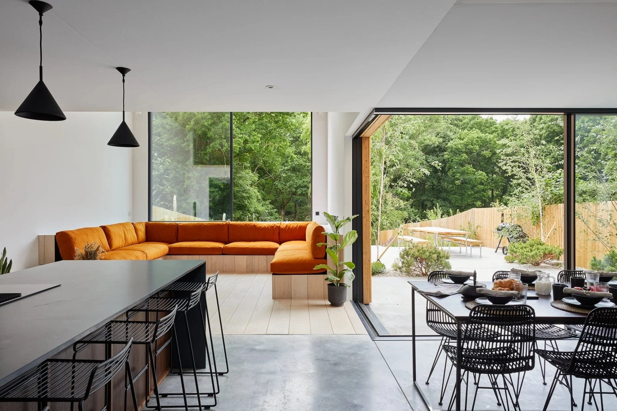 Modern open-concept living and dining area with large sliding glass doors opening to a backyard with trees and outdoor furniture. Indoor features include an orange sectional sofa, black pendant lights, a dining table with black chairs, and minimal decor.