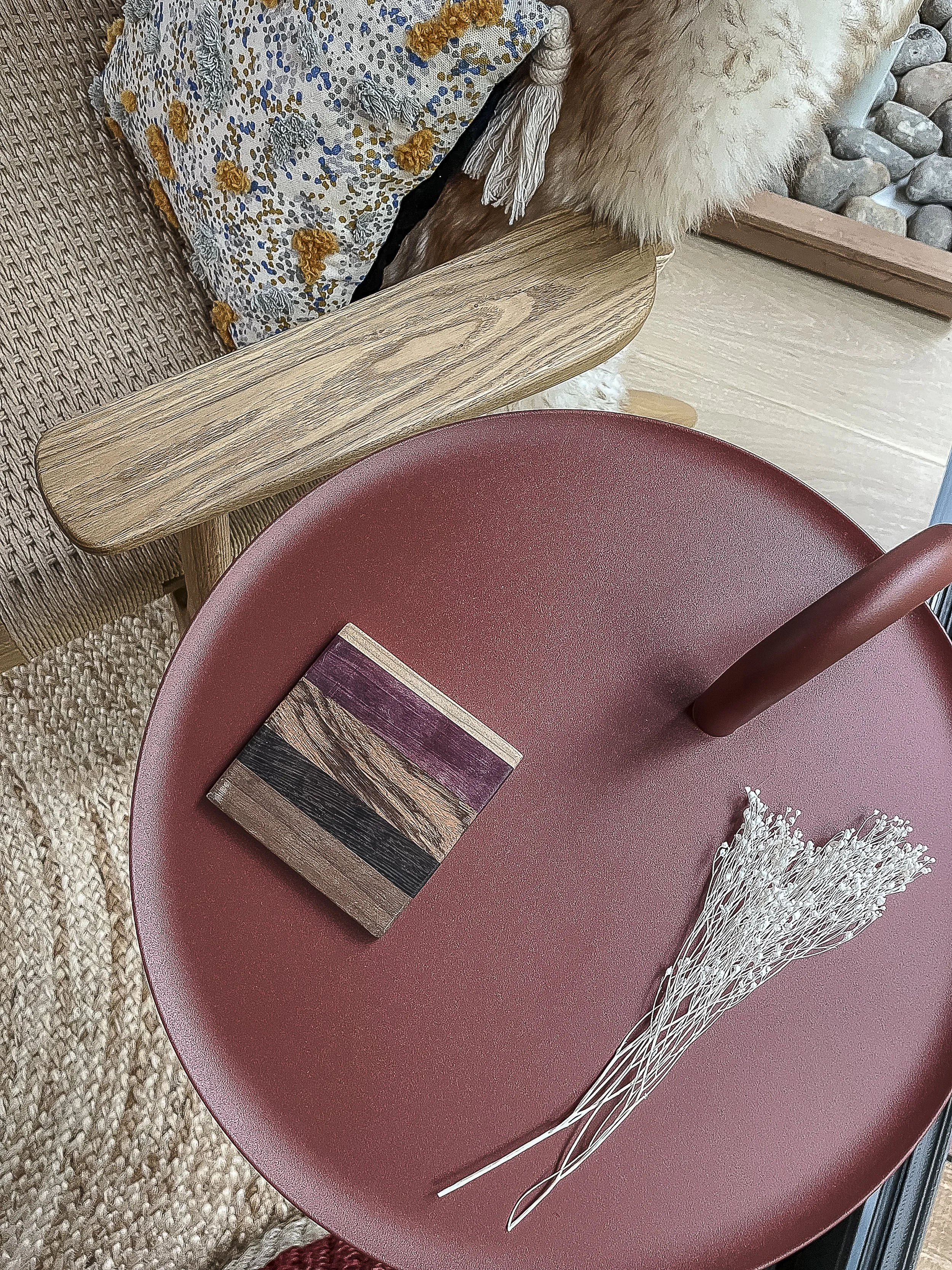 A red circular table with a small wooden block and a bundle of white dried flowers on it. Part of a wooden chair and a colorful pillow with yellow and blue patterns are visible, along with a beige textured rug and a section of wooden flooring.