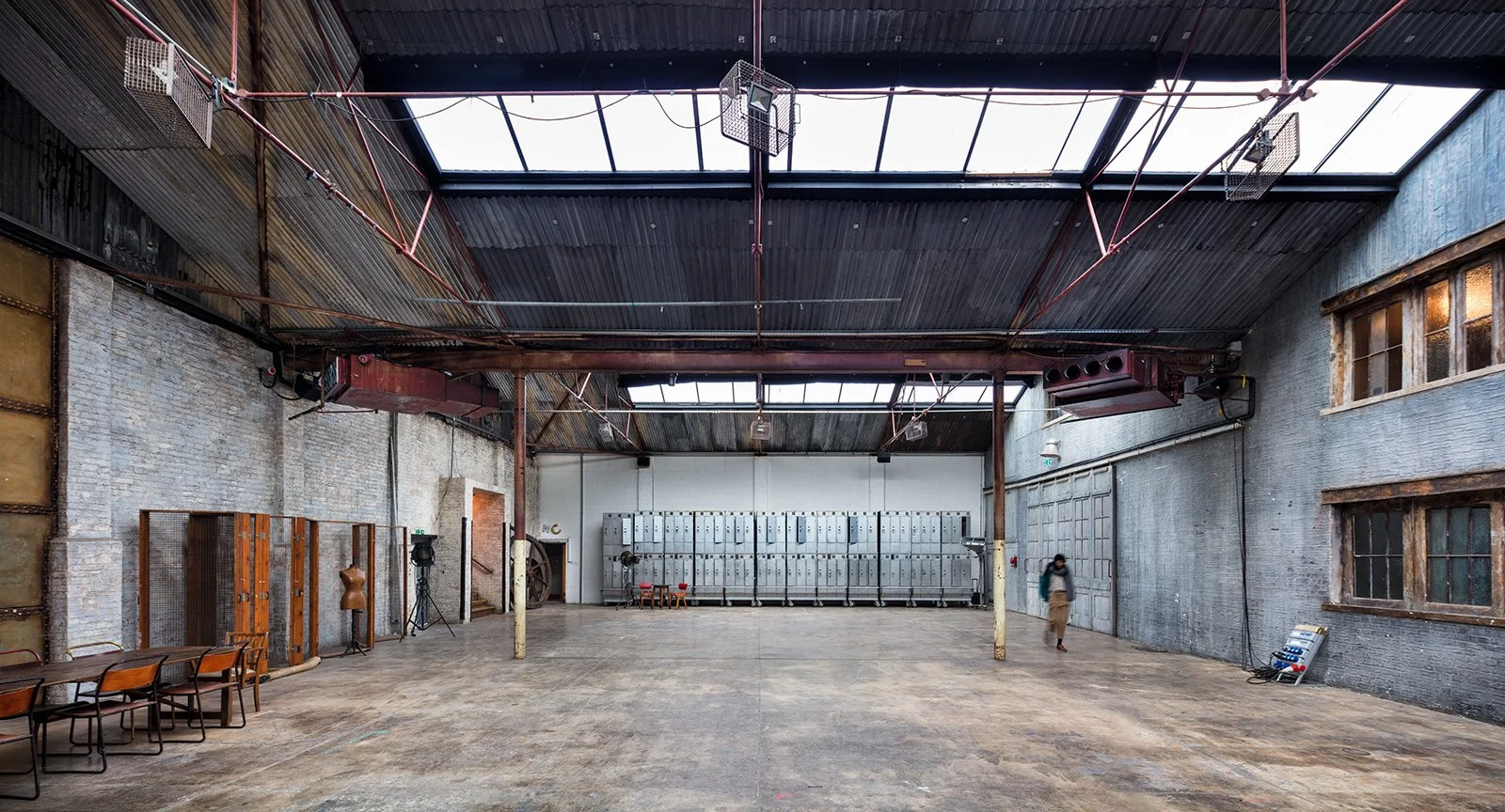 Interior of an industrial building with a high metal roof, skylights, lockers, a person walking, chairs, and equipment.