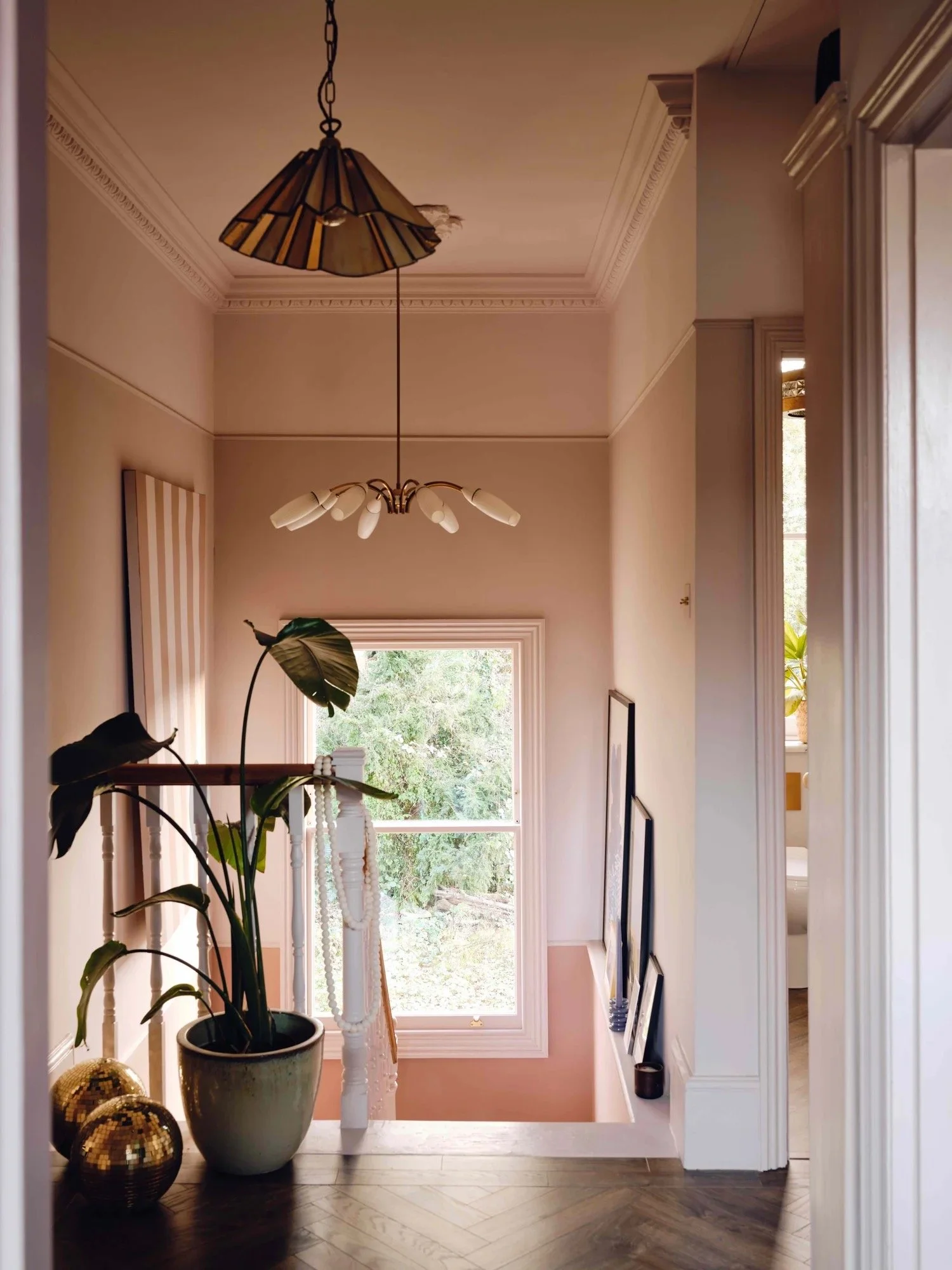 Interior staircase landing with a large window, framed photos on the right wall, a large potted plant, and decorative gold spheres on the floor, illuminated by a ceiling light fixture.