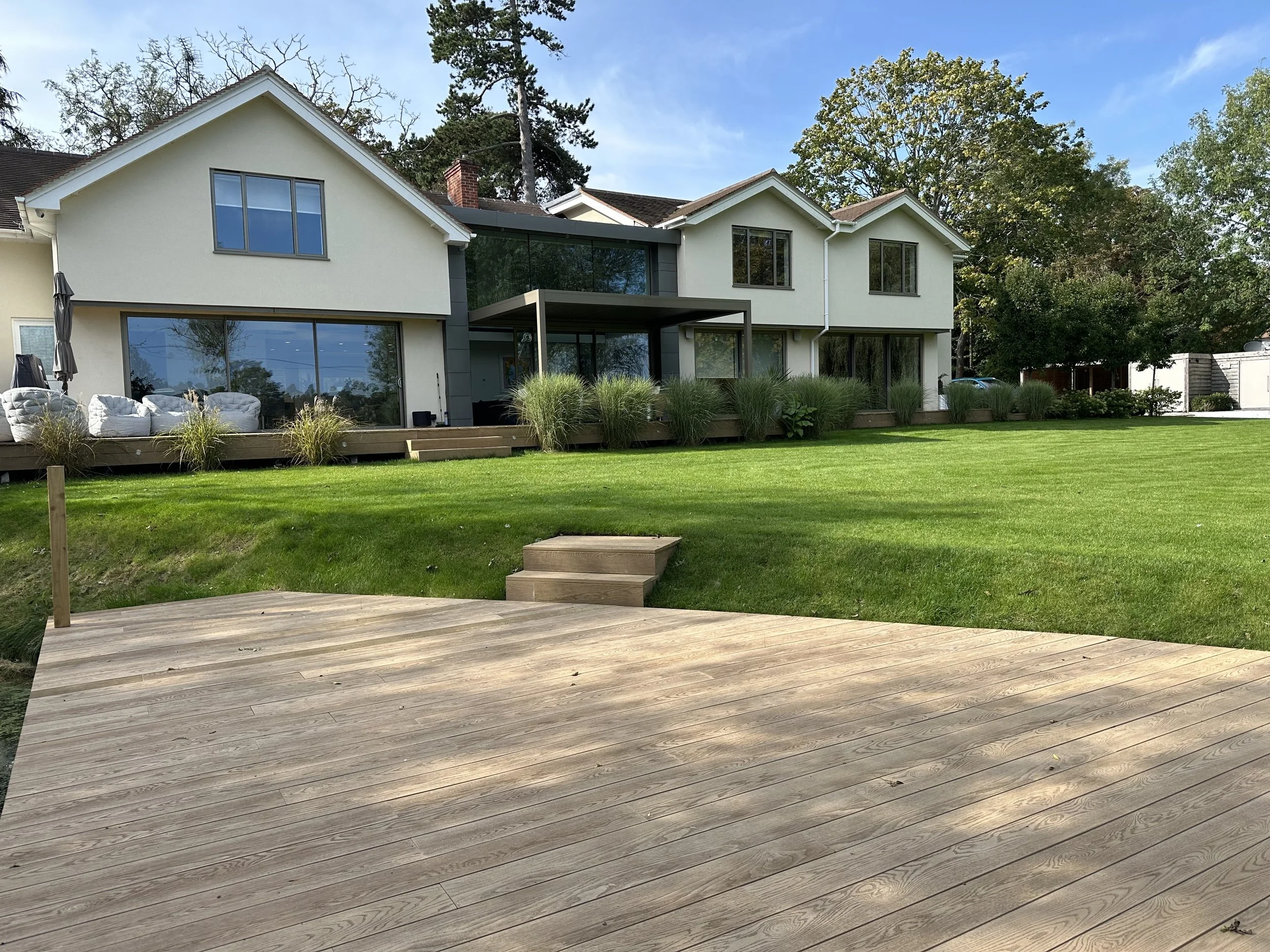 Modern two-story house with large glass windows, a wooden deck, and well-maintained lawn and garden with trees in the background.