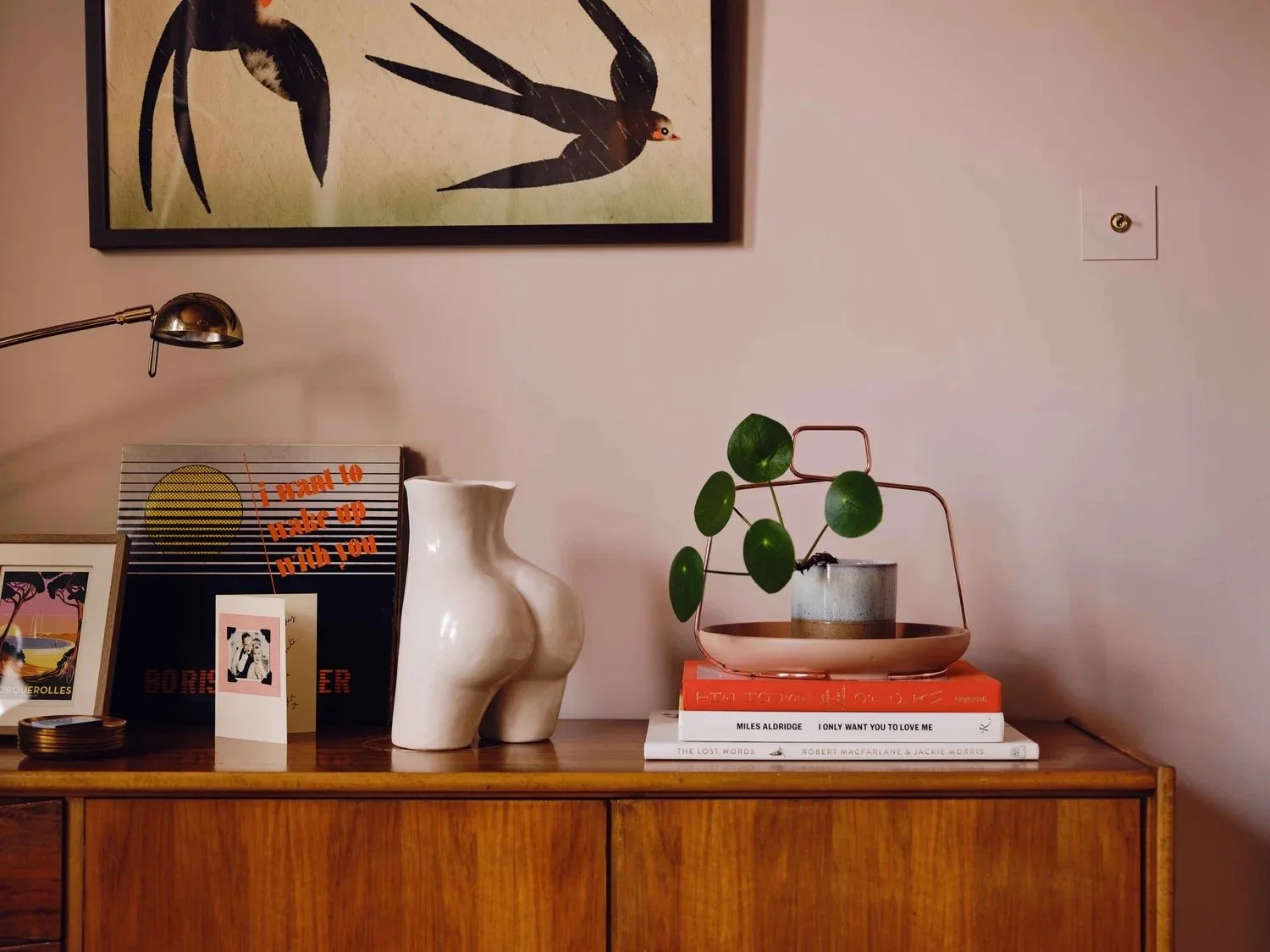 Decorative tabletop with art, books, and sculptures, including a ceramic abstract female torso, a framed photograph, a potted plant with green round leaves, a stack of books, and a modern gold desk lamp, against a pink wall with a framed art piece ab