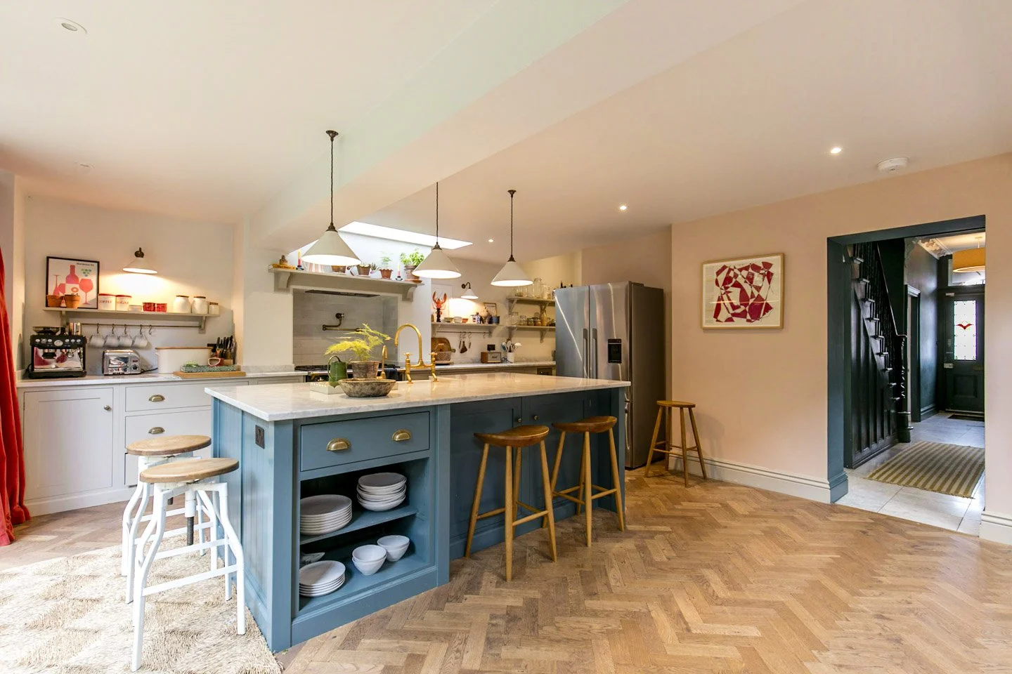 Bright open kitchen with a large blue island, white countertops, wooden barstools, pendant lights, stainless steel refrigerator, open shelving, and a doorway leading to entryway with striped rug.