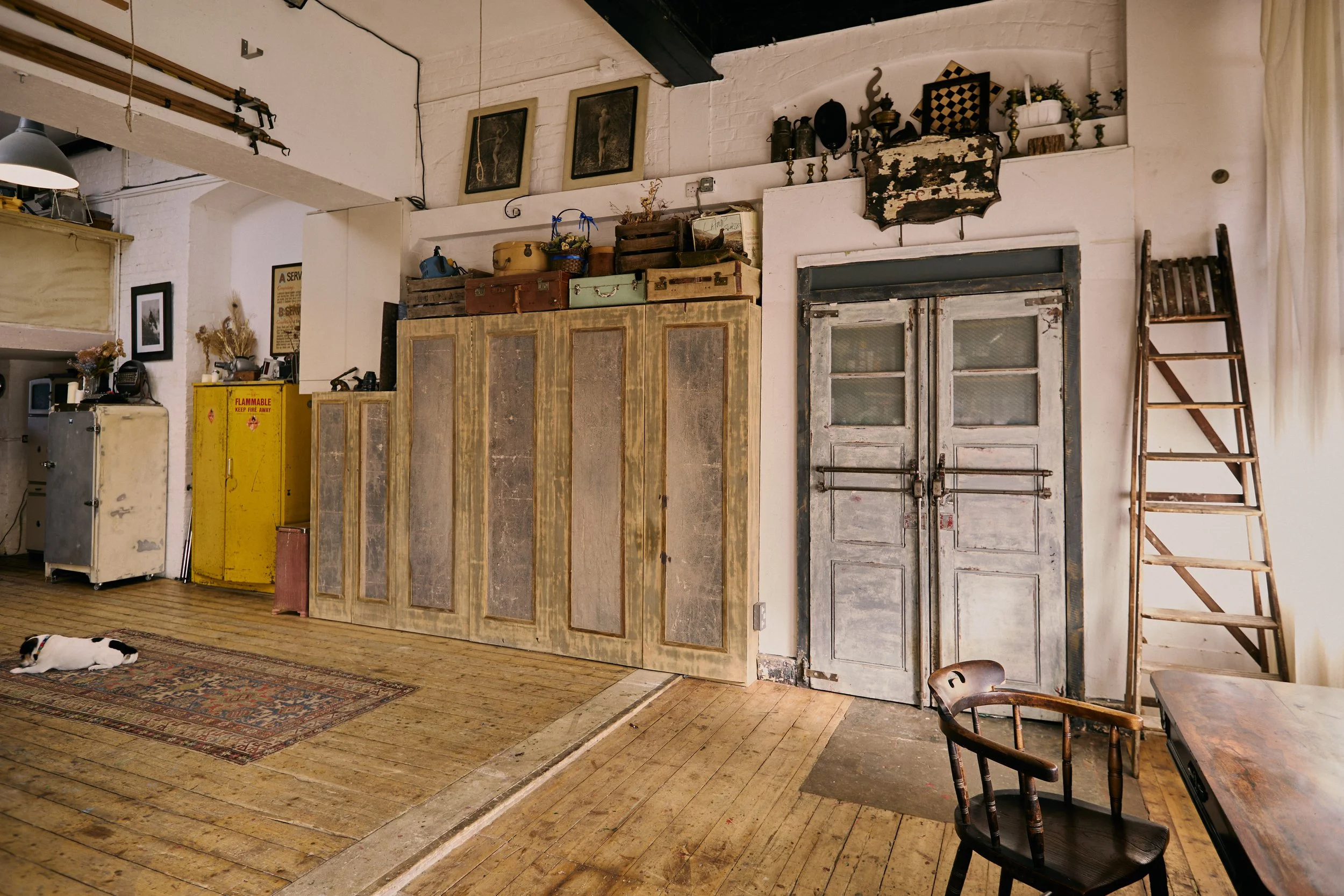 Interior of a rustic room with vintage wooden furniture, a closed pair of metal double doors, an old ladder, a patterned rug on wooden floor, and a small dog lying on the rug.