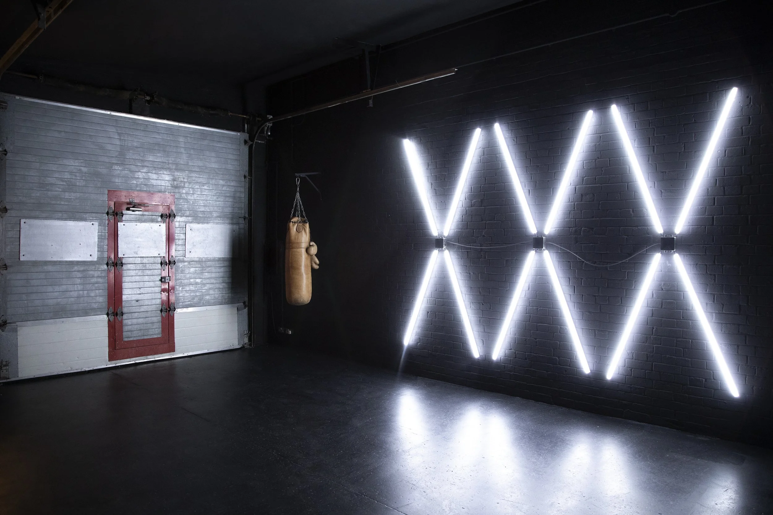 A black wall with bright white neon light installations arranged in a zigzag pattern, a brown punching bag hanging on the wall, and a metal garage door with a red and gray frame.