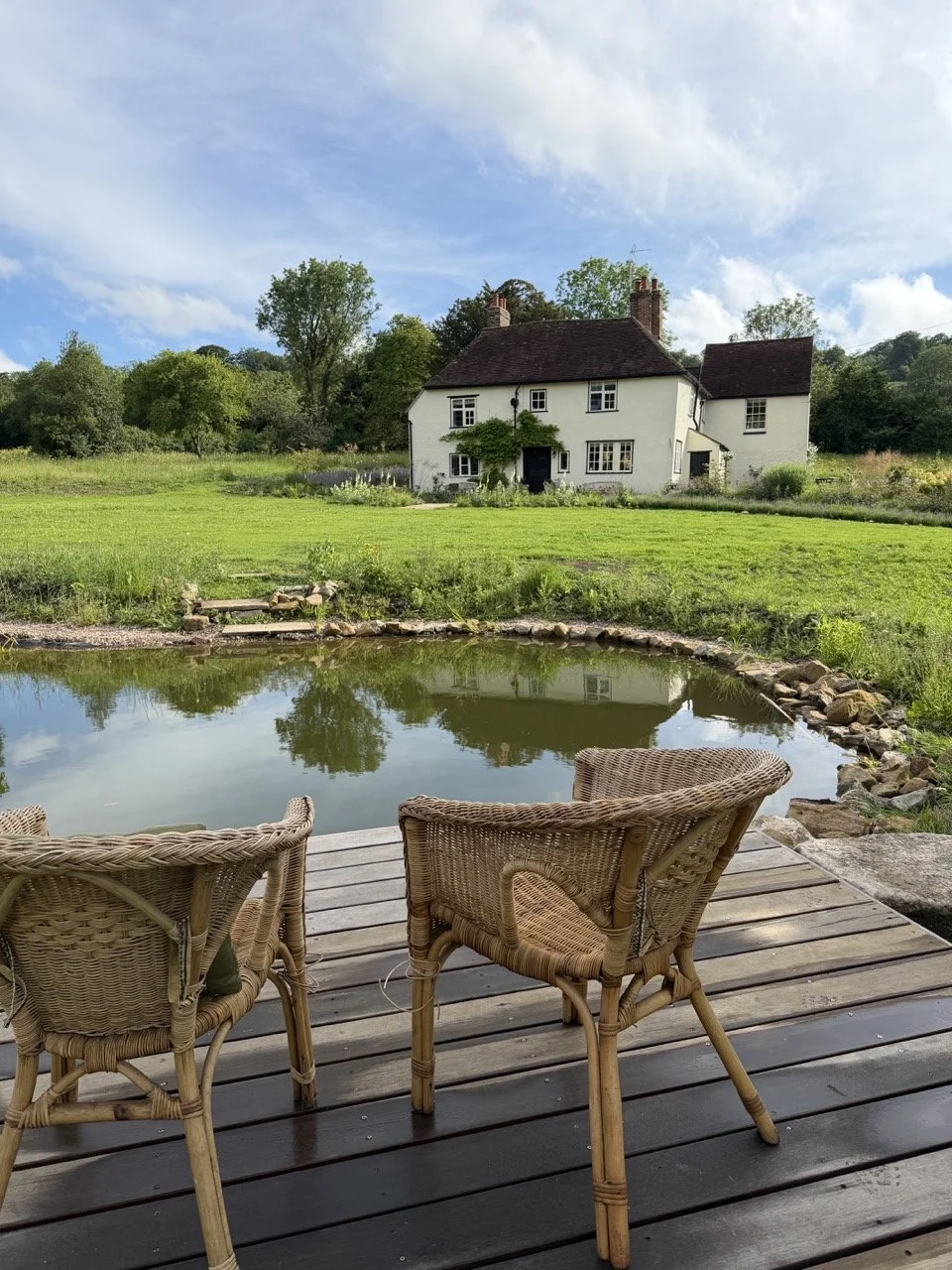 Wicker outdoor chairs on a wooden deck overlooking a pond with a white house and green trees in the background under a partly cloudy sky.
