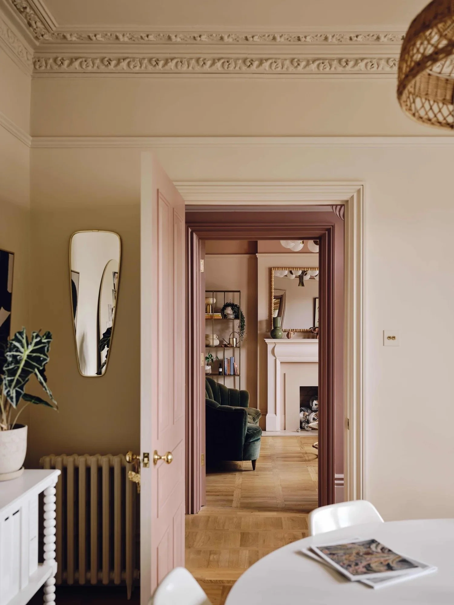 View through open doorways into a living room with a fireplace, bookshelf, and green armchair. A round dining table with magazines is in the foreground.