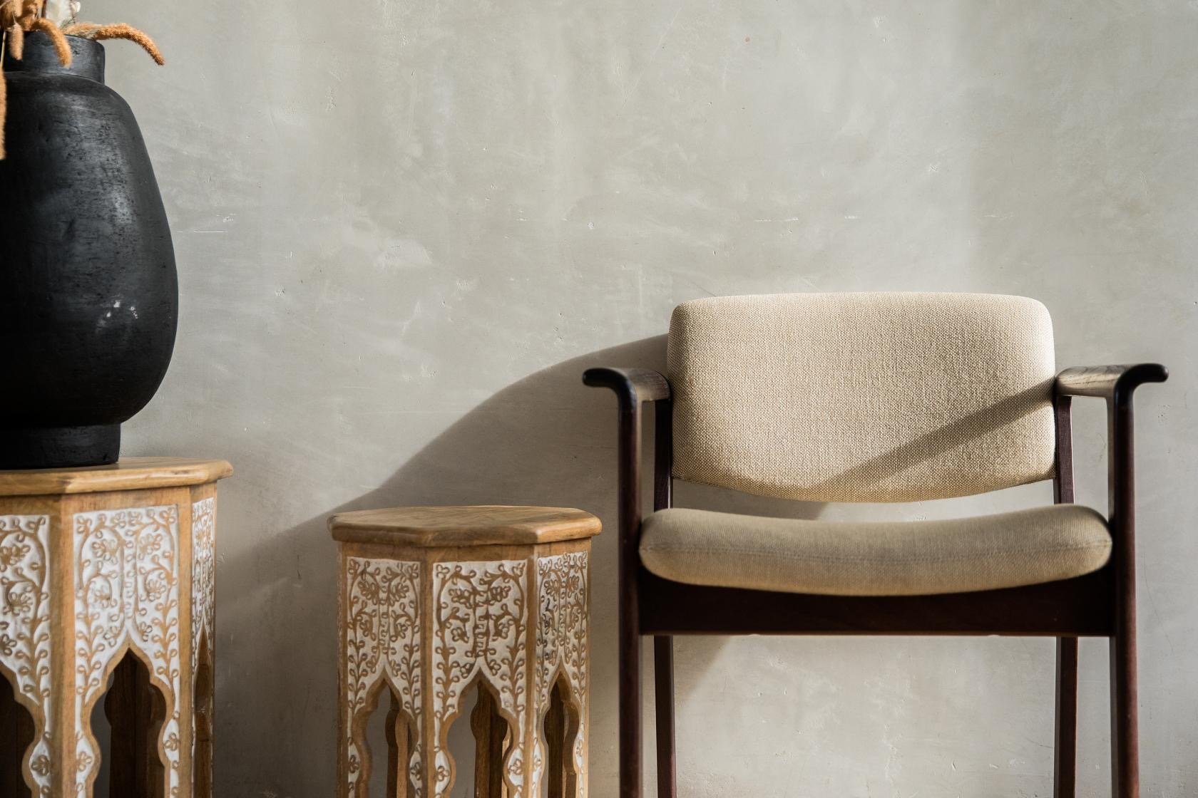 A beige cushioned chair with dark wooden arms and legs, flanked by two ornate white and light brown carved side tables, with a large black vase on a wooden table to the left, against a plain, light-colored wall.