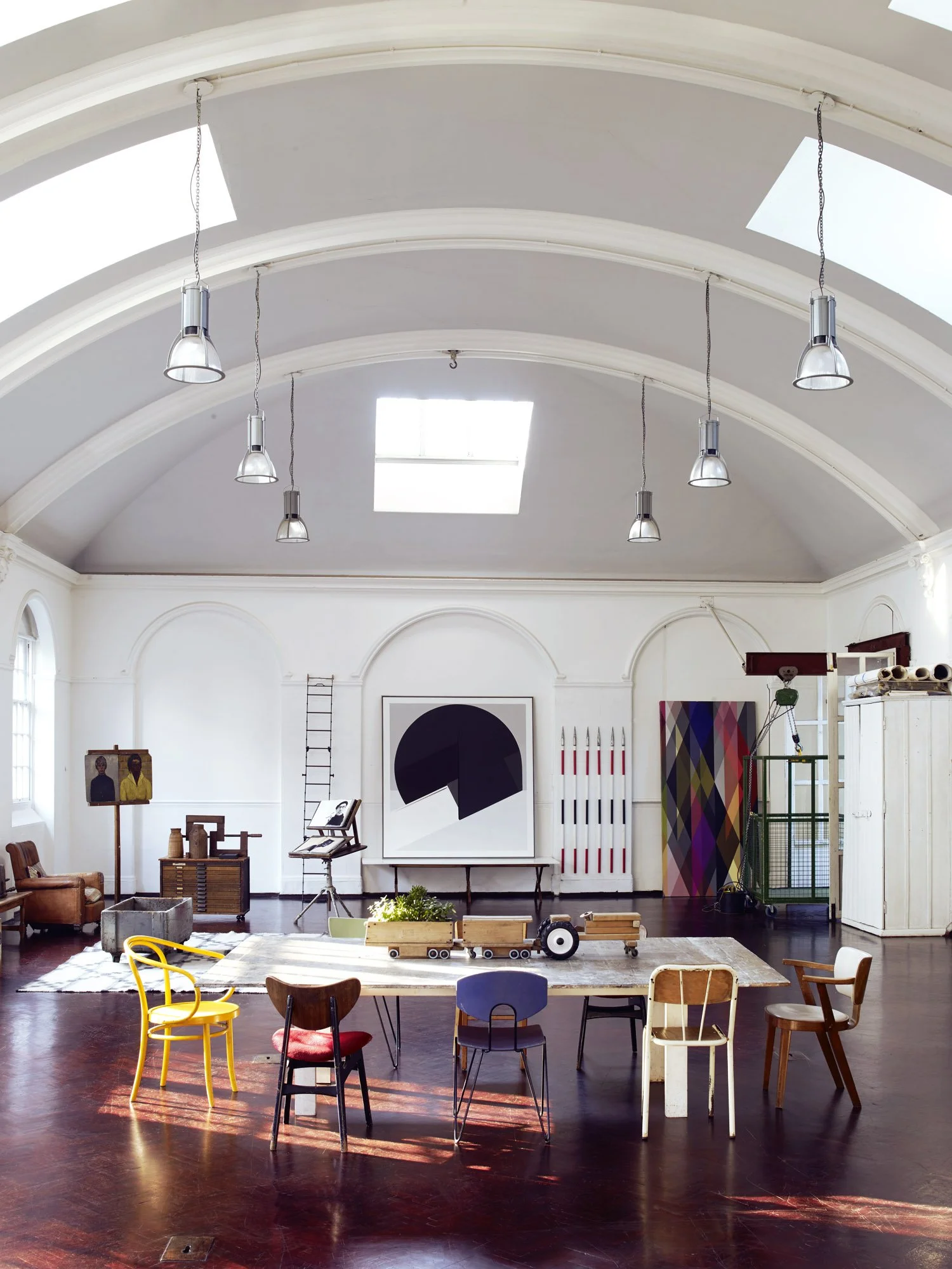 Interior of a spacious art studio or gallery with high arched ceiling, skylights, and various chairs around a central table. Artwork and art supplies are visible in the background.