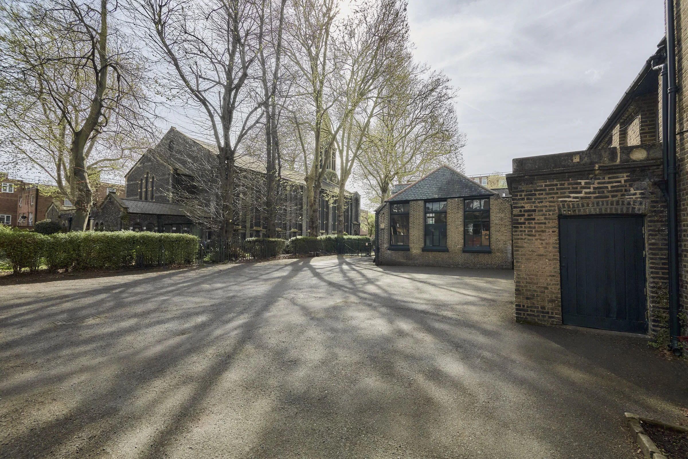 An outdoor scene with a gravel open area, leafless trees casting shadows, a stone building with large windows in the background, and brick buildings on the right side.