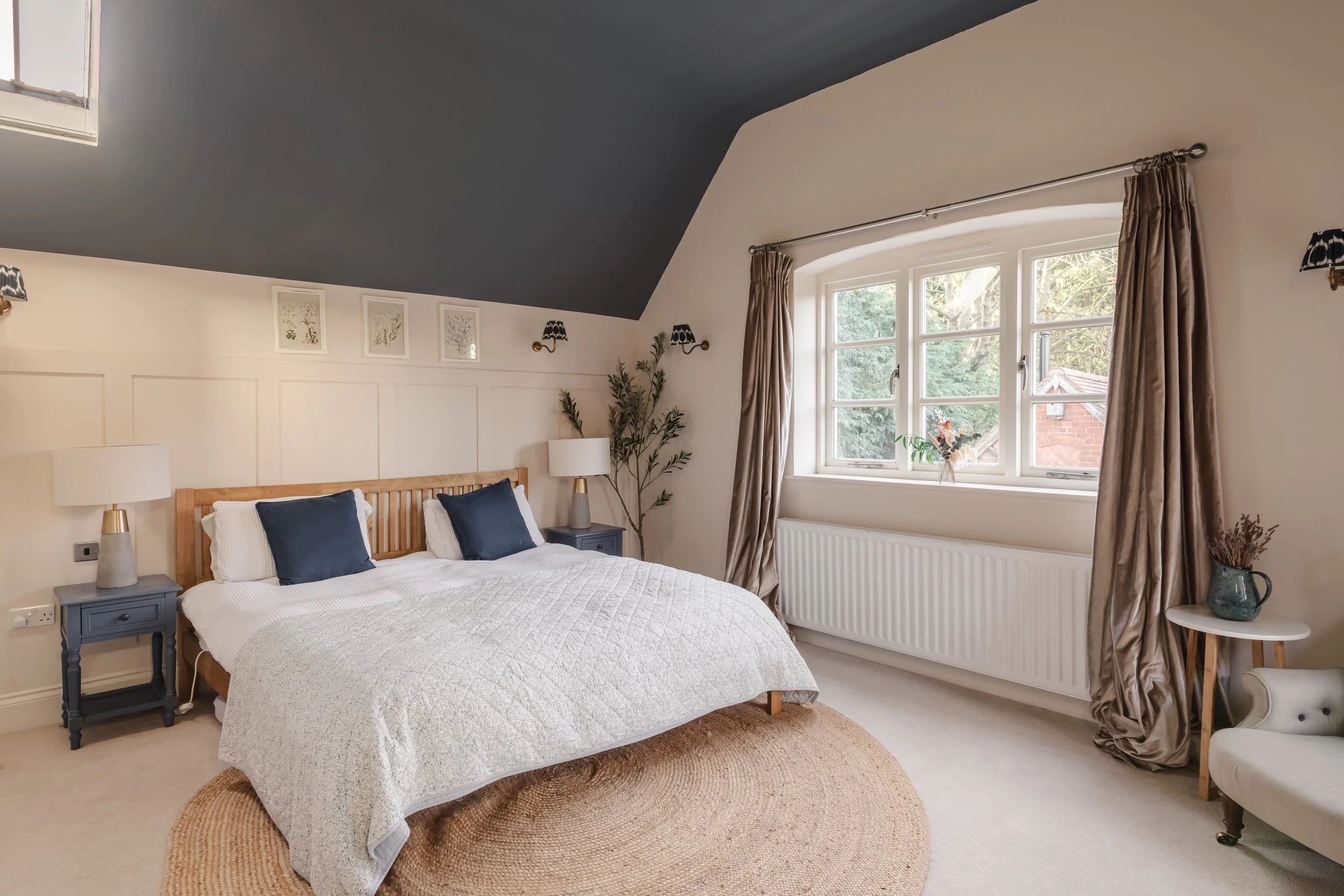 A cozy bedroom with a large bed, dark blue pillows, white bedding, two blue nightstands with lamps, a window with beige curtains, a decorative plant, and a round jute rug.