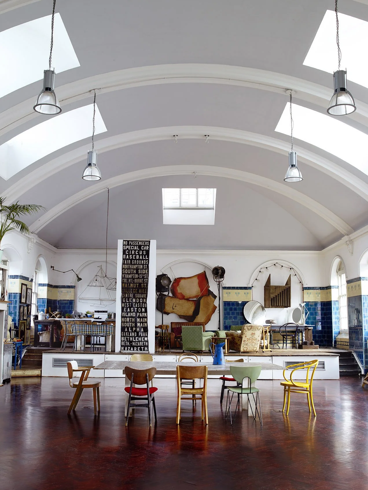 Interior of a spacious room with a high vaulted ceiling, skylights, hanging lamps, and eclectic furniture including chairs, sofas, and a table. Walls are white with blue tile accents, and there's a black and white sign with list of locations or names