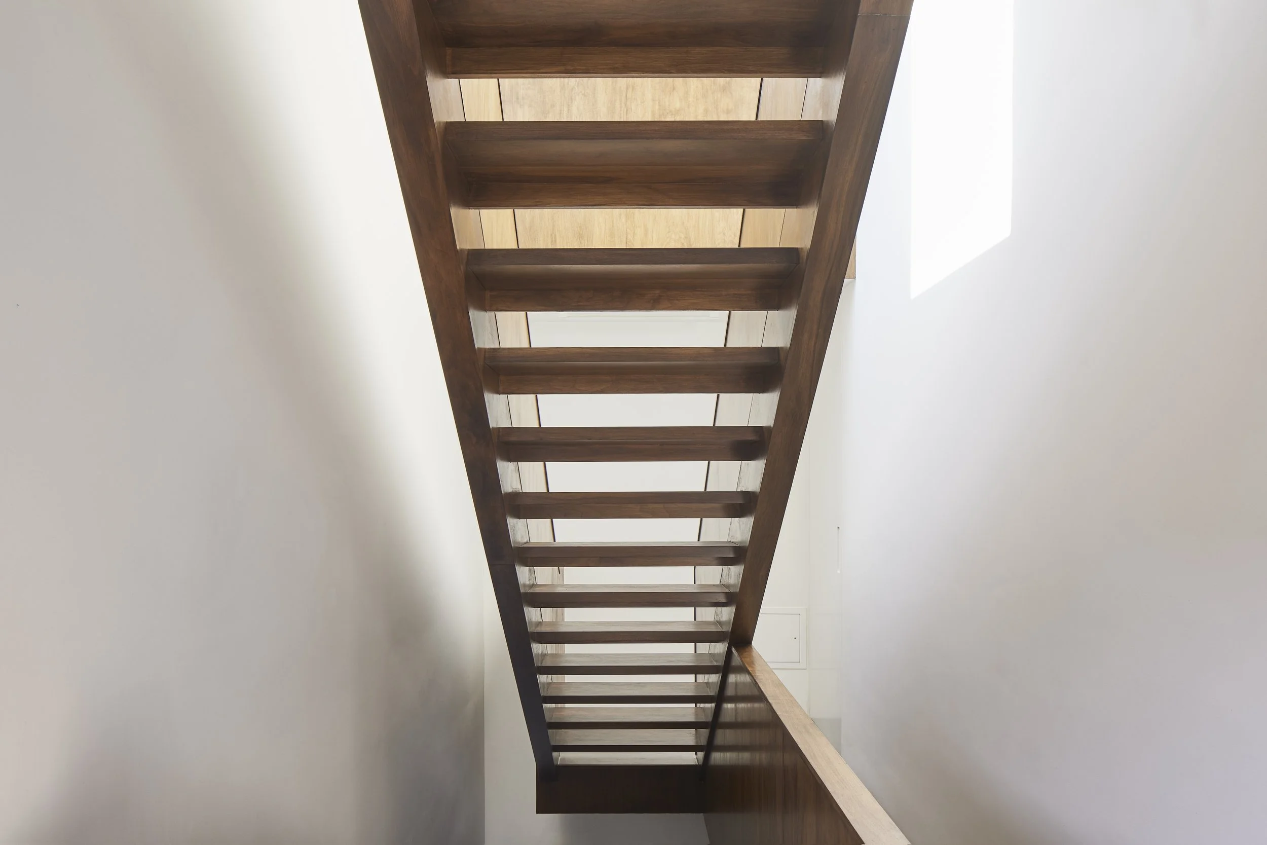 Looking up at a wooden staircase with open risers, built against a white wall and illuminated by natural light from a window.