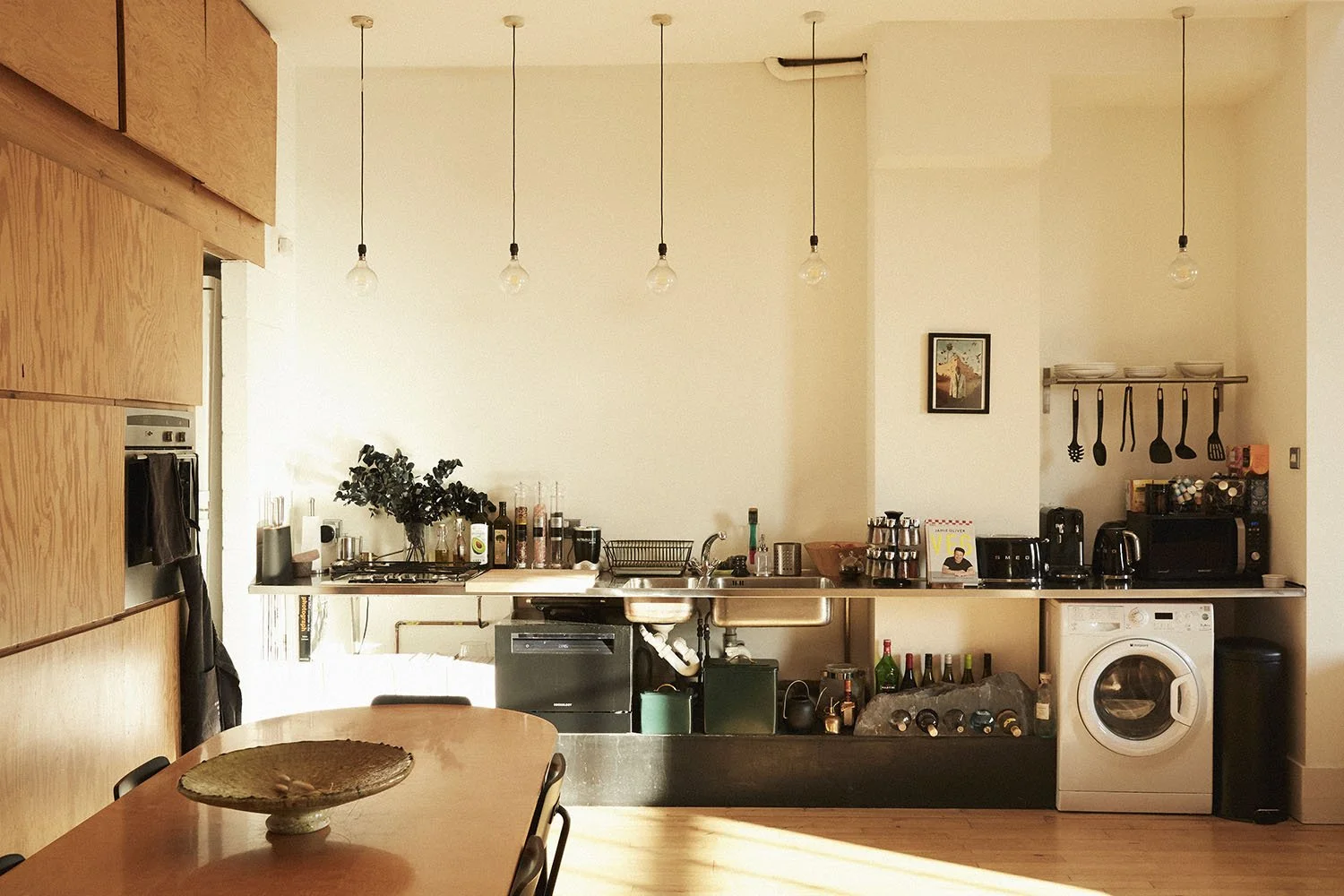 Modern kitchen with wooden cabinets, hanging light bulbs, a stainless steel sink, various bottles and kitchenware on the counter, and a washing machine. A dining table with a decorative plate is in the foreground.